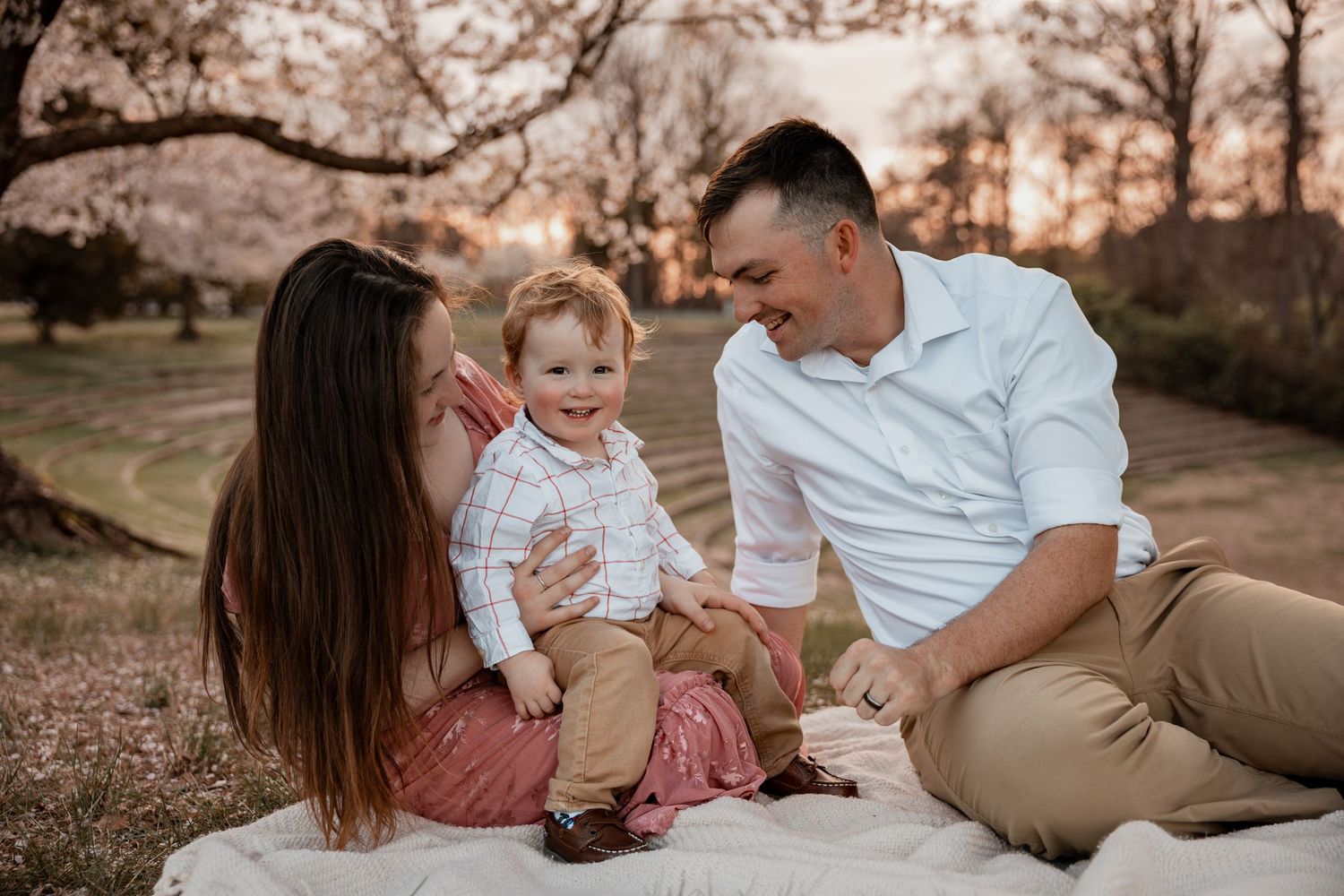 A happy young family sits on a blanket outdoors at sunset, with parents smiling at their cheerful baby boy.
