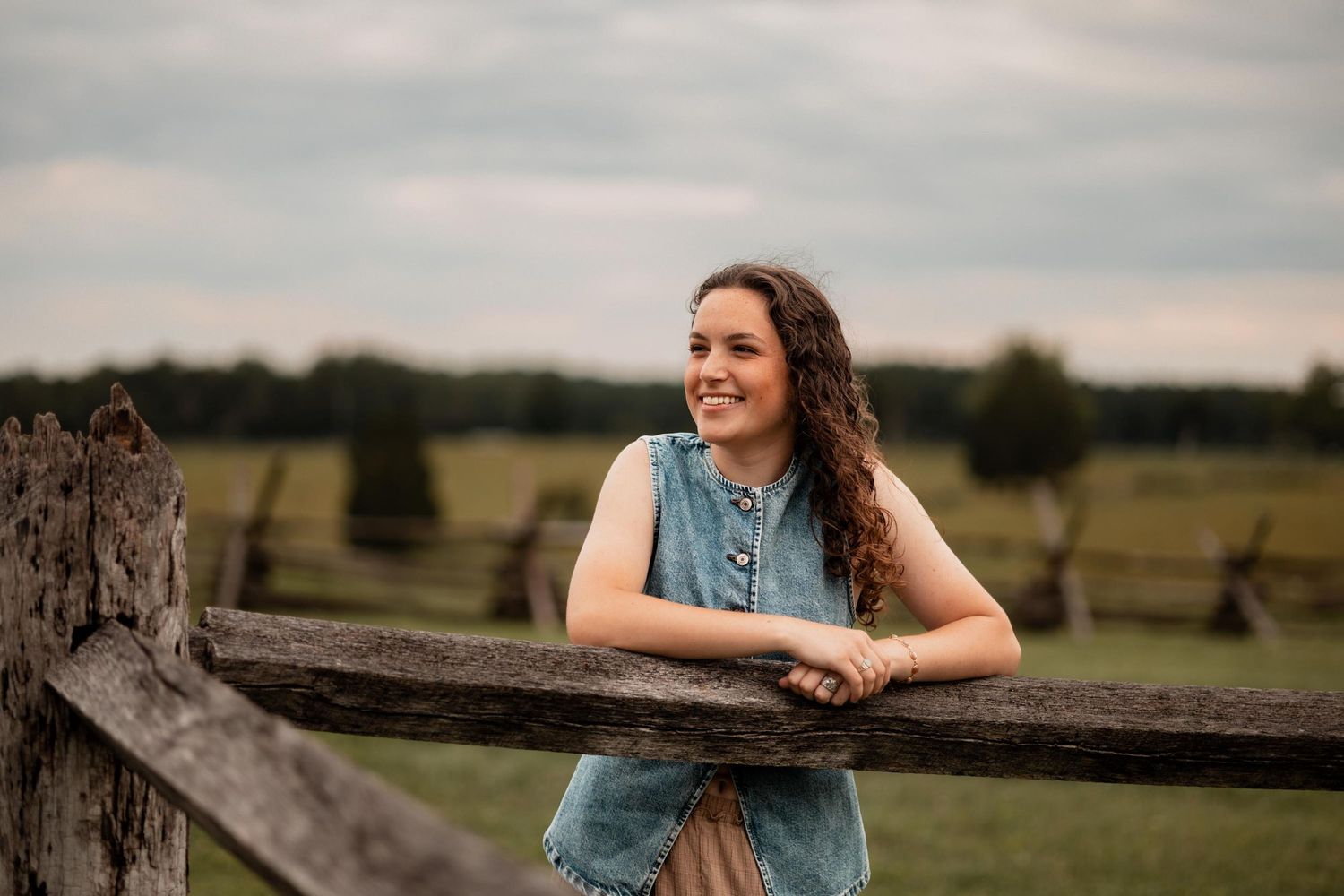 Young woman in denim vest smiling while leaning on rustic wooden fence in green countryside setting.
