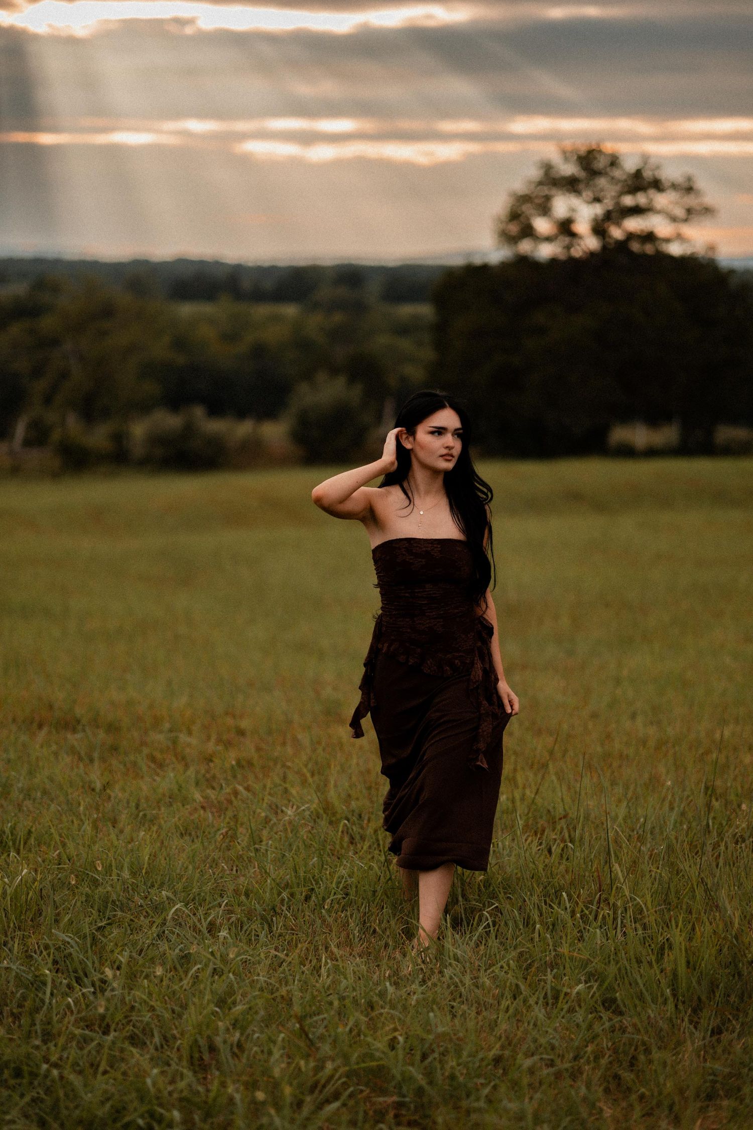 Woman in dark strapless dress walks through green field at sunset with dramatic rays of light.