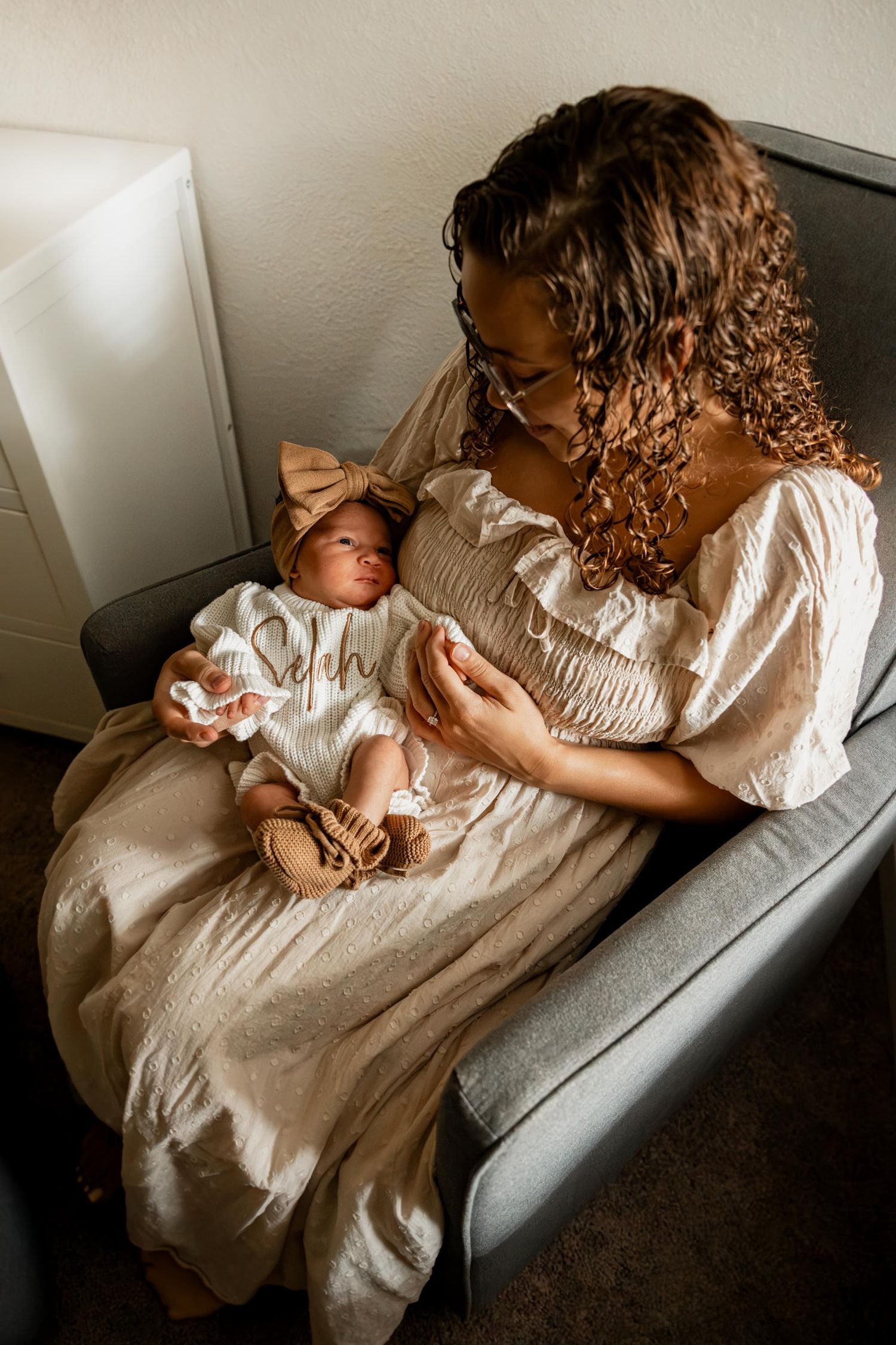A curly-haired mother in a cream dress tenderly holds her newborn baby on a gray sofa.