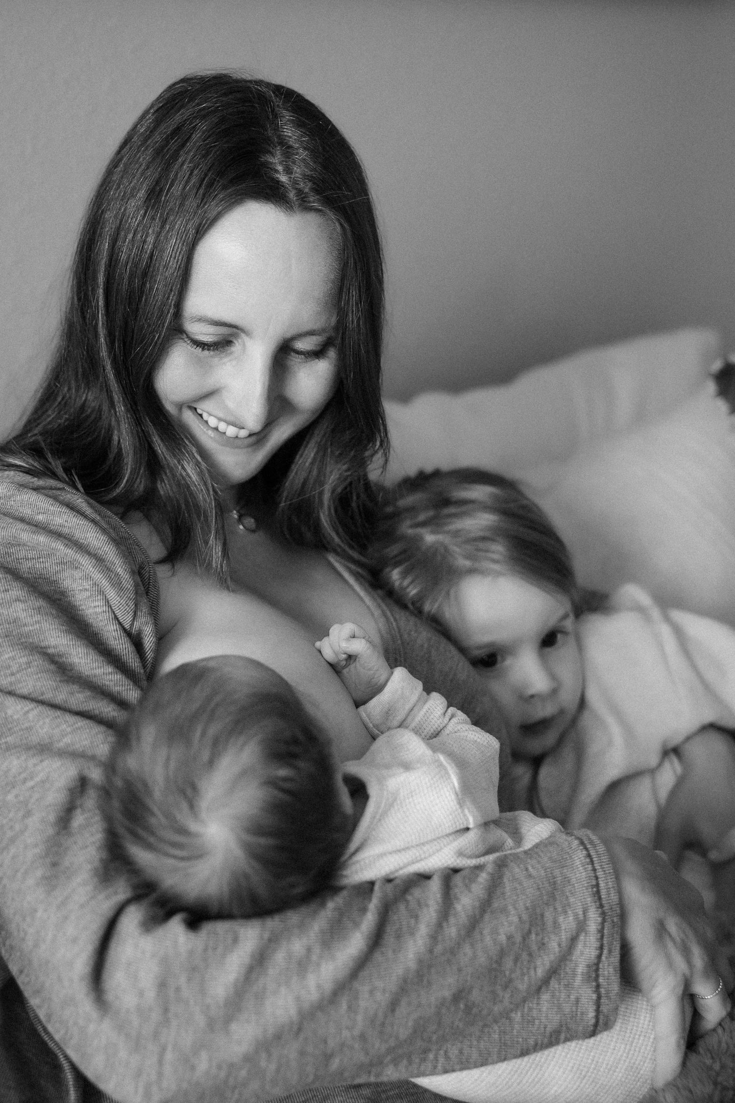 Black and white photo of a smiling mother breastfeeding newborn with toddler beside her.