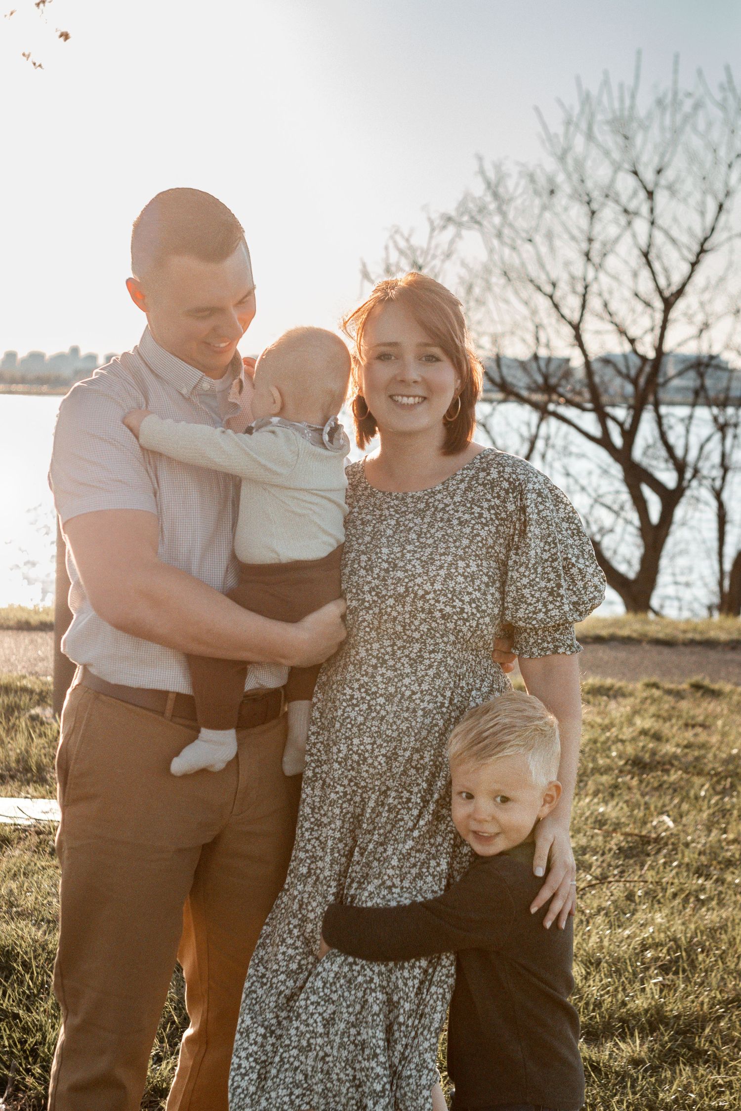 A family enjoys a sunny afternoon outdoors together under bare trees on a grassy hill by the water.