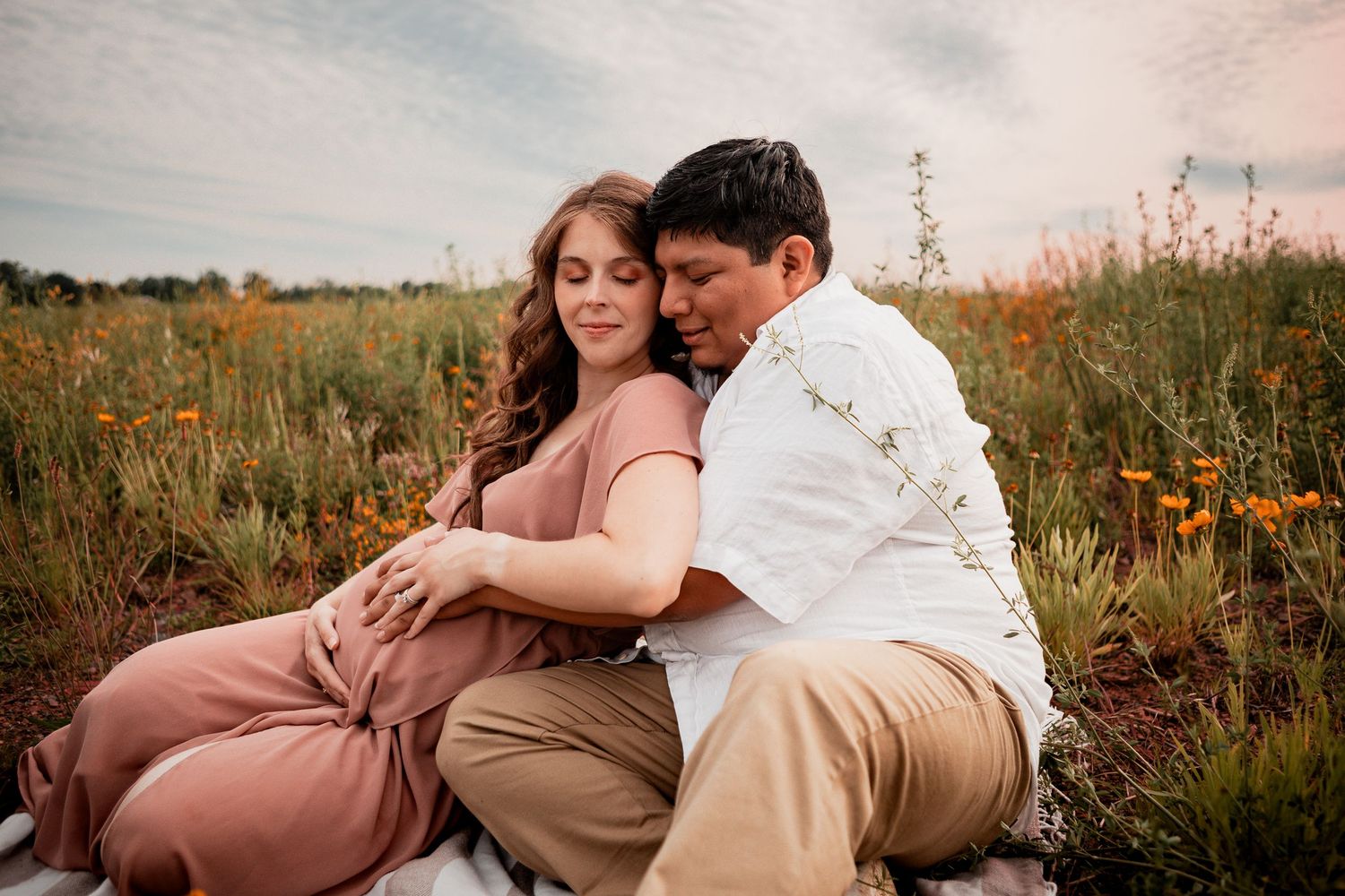 Romantic pair embrace tenderly in a sunlit meadow with tall grass and wildflowers during golden hour.