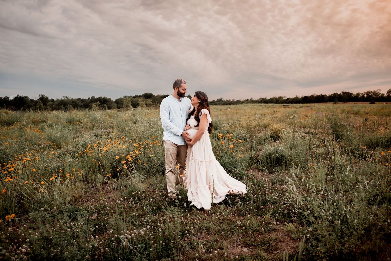 A couple in white wedding attire embraces in a scenic prairie field during sunset.