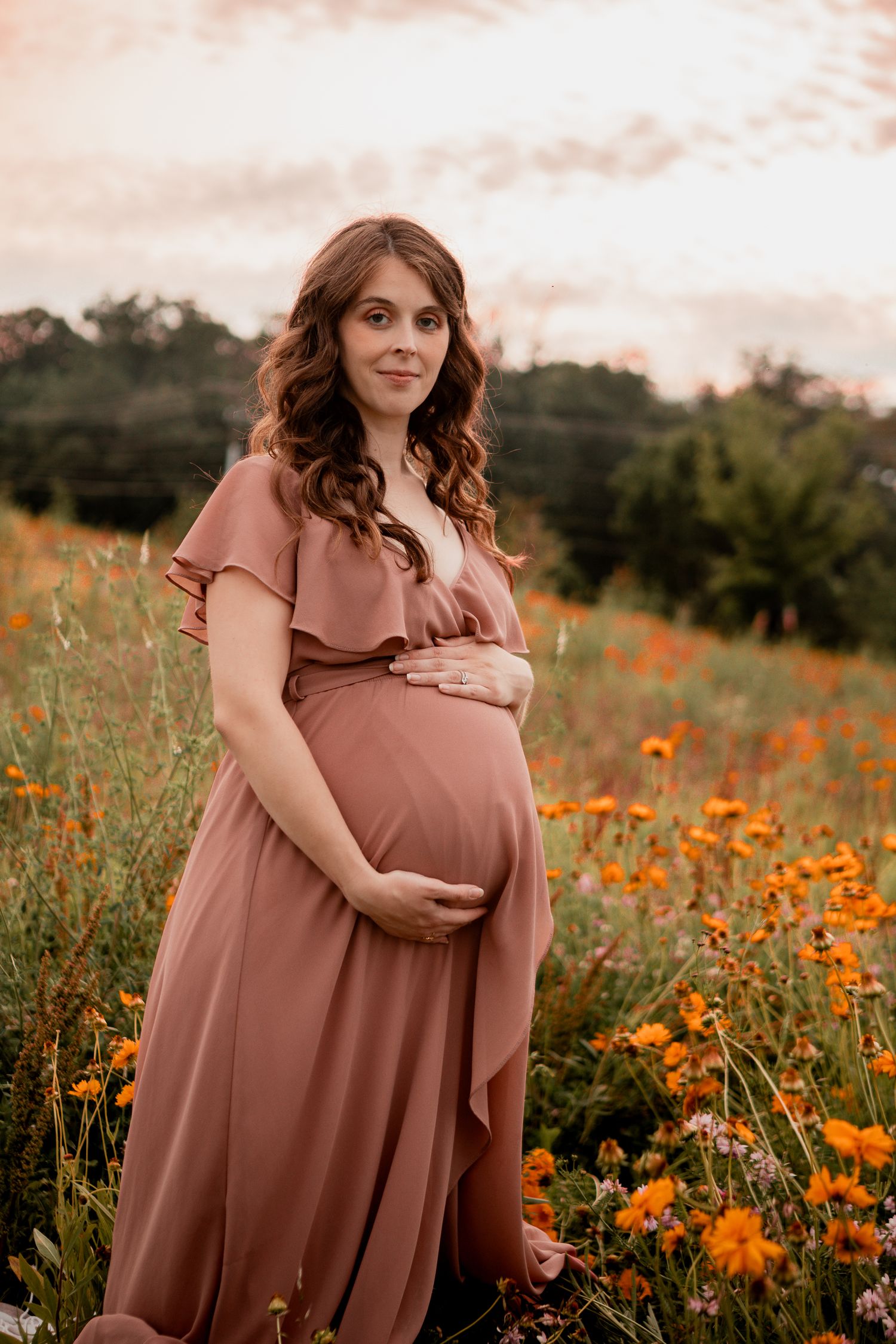 A woman in a dusty pink dress cradles her pregnant belly while standing in a field of orange poppies at sunset.