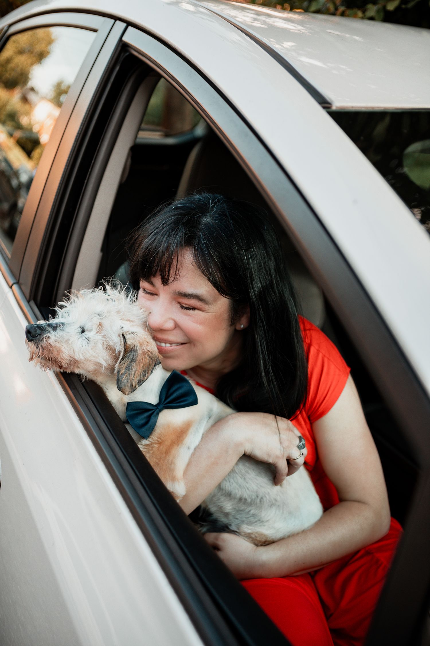 A small white dog and a person in a red top share a sweet moment inside a car.