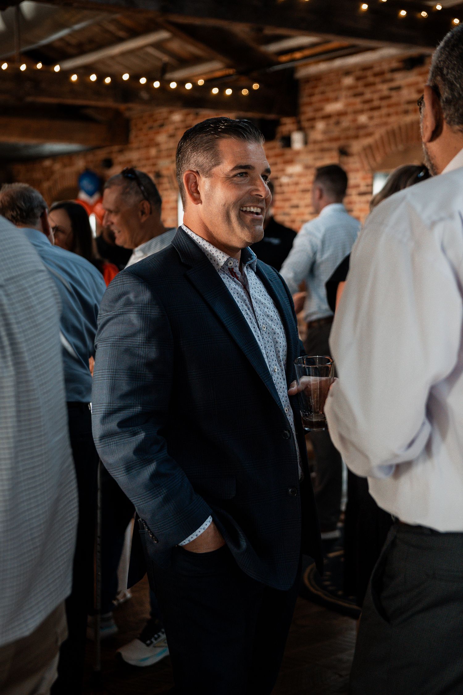 Business professionals in suits mingle and socialize at an event with exposed brick walls and string lights.
