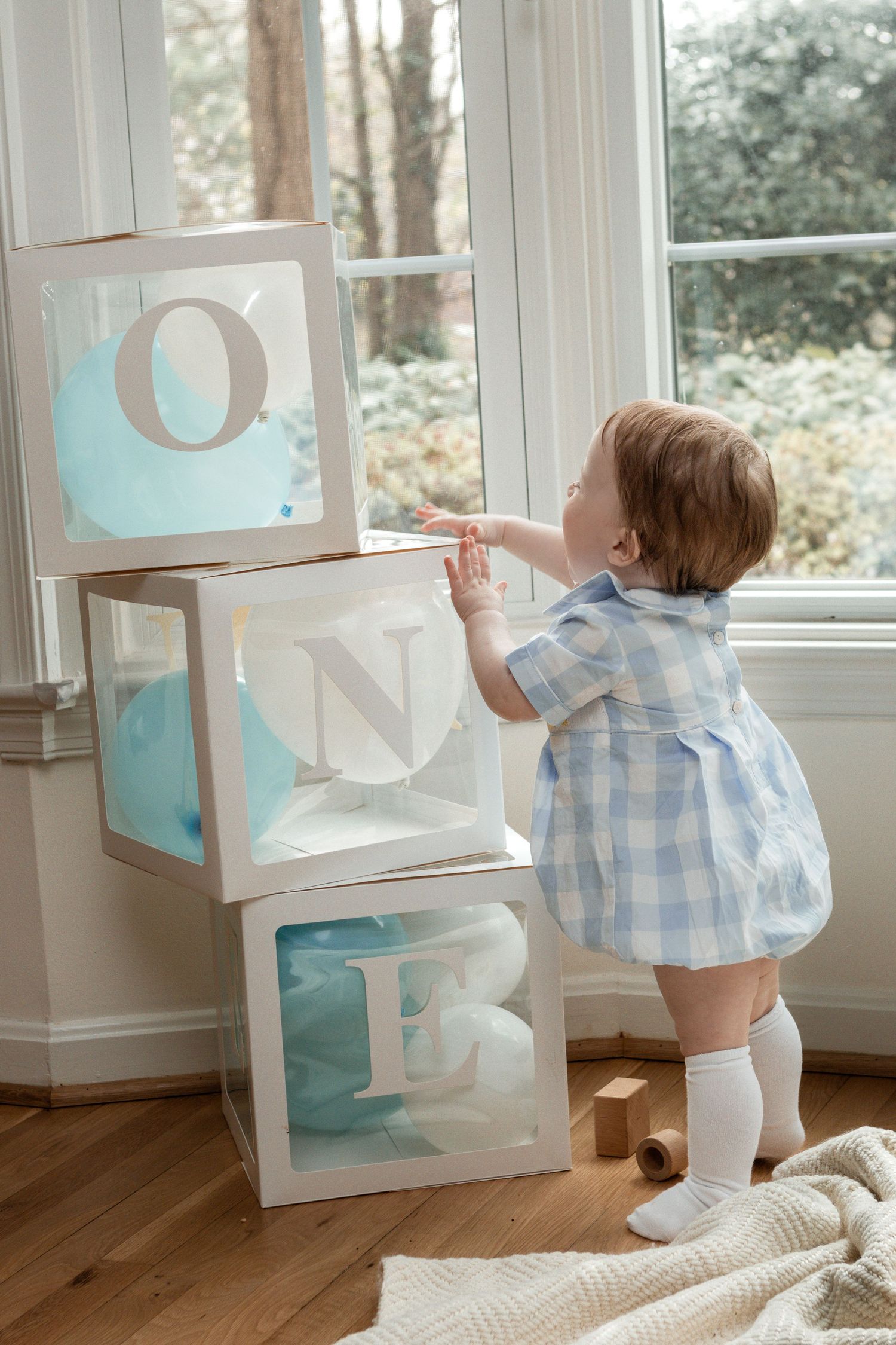 Large decorative baby blocks spelling ONE displayed near a window as a small child in blue gingham reaches up to touch them.