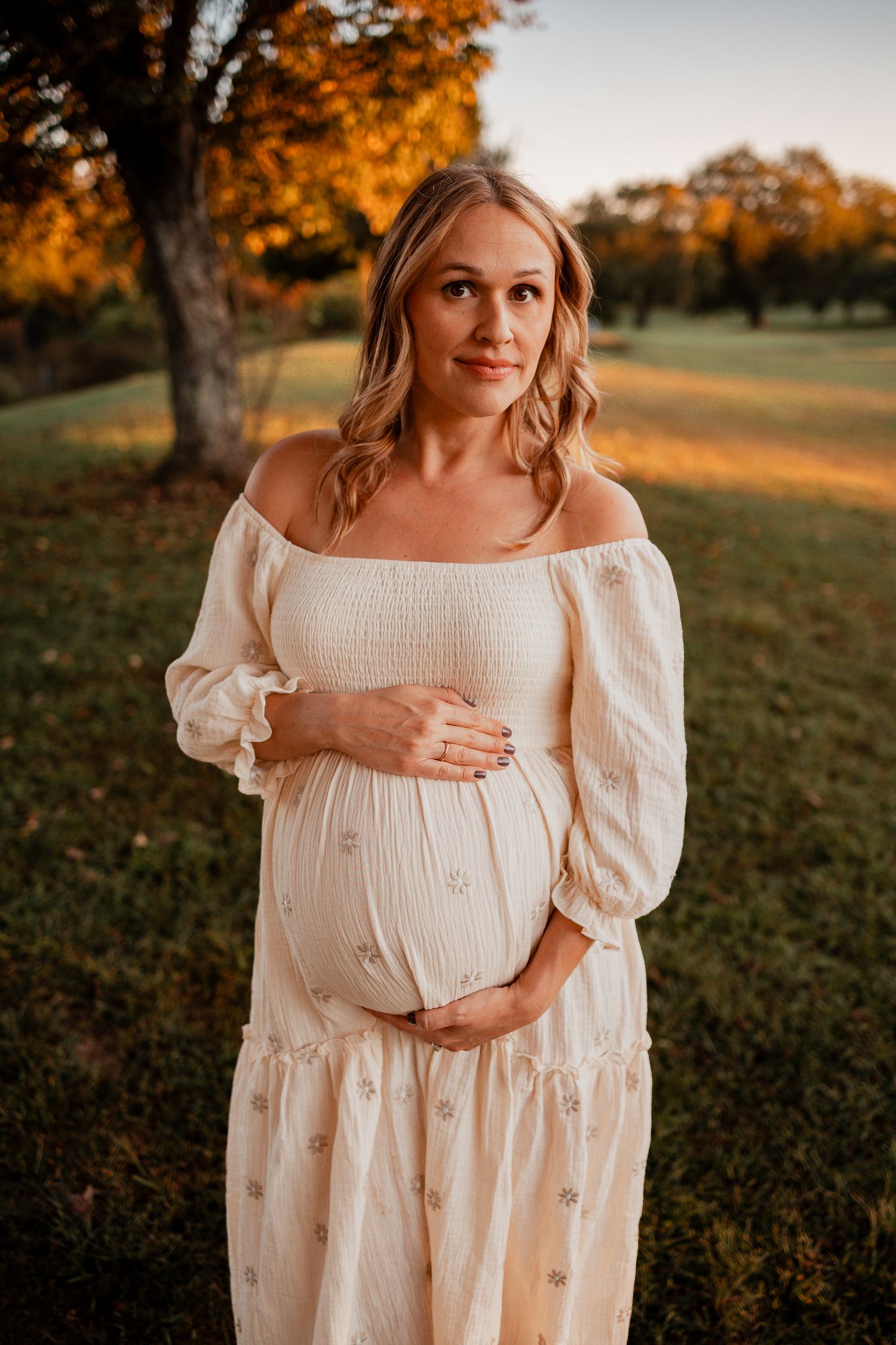 Pregnant woman in white off-shoulder dress cradling baby bump outdoors during golden hour sunset.