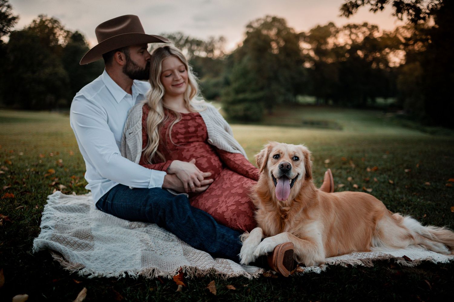 A couple relaxes on a picnic blanket with their golden retriever at sunset.