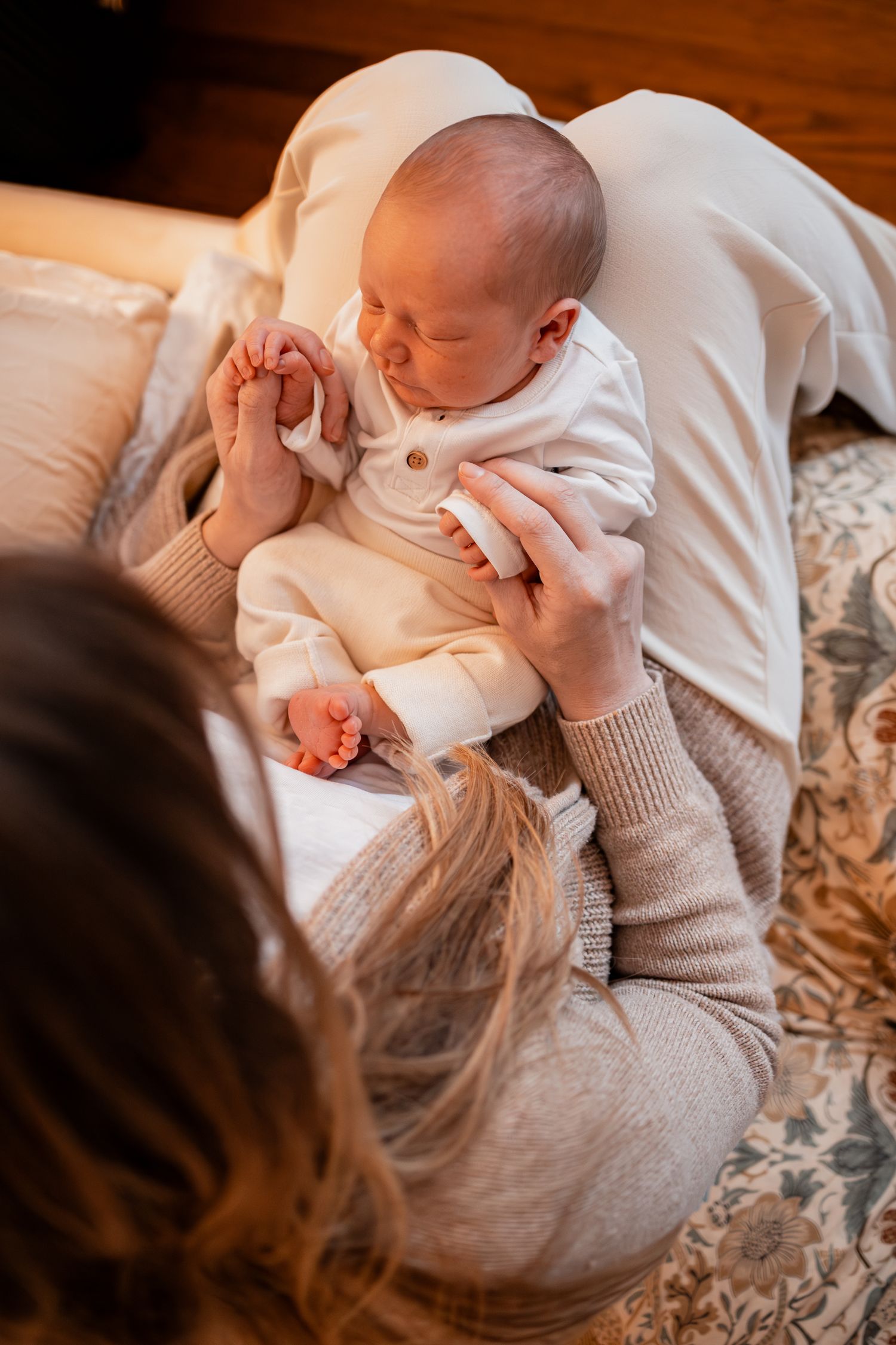 A tender moment with a baby in white clothing being held on a patterned bedspread in warm lighting.