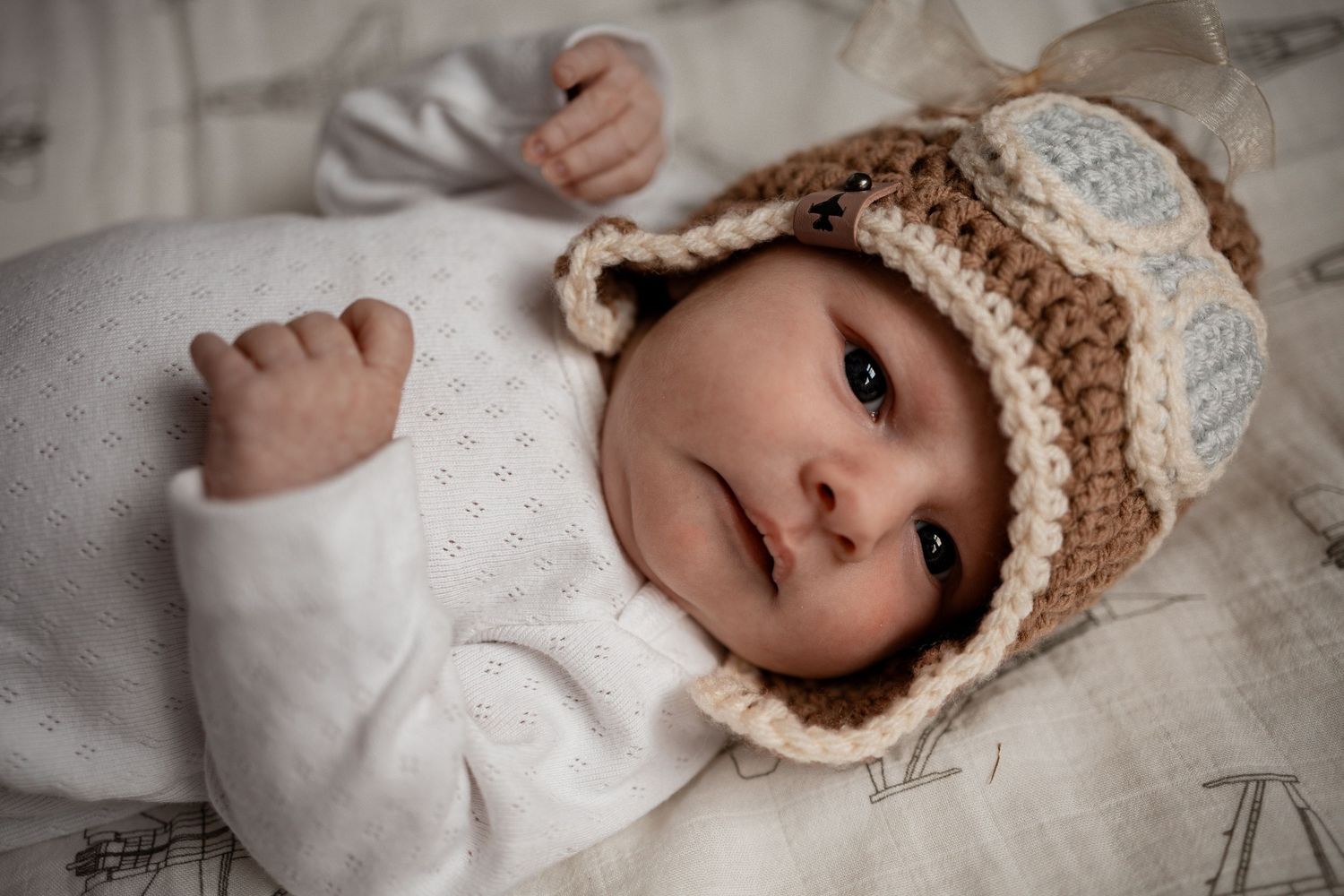 Baby wearing a knit bear hat lies on white bedding with floral pattern details.