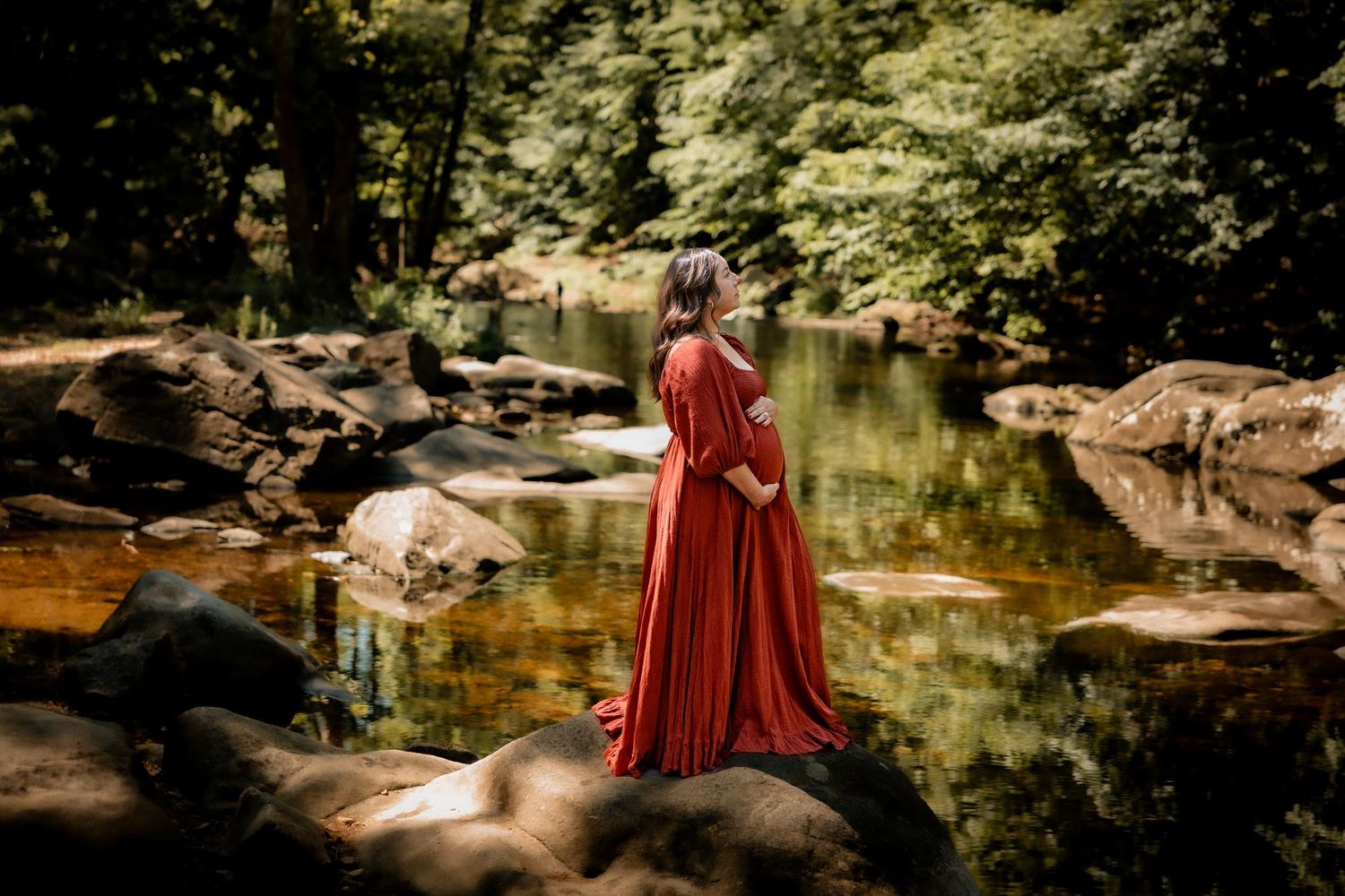 Pregnant woman in red gown standing on rocks by a reflective forest stream during a maternity photo session.