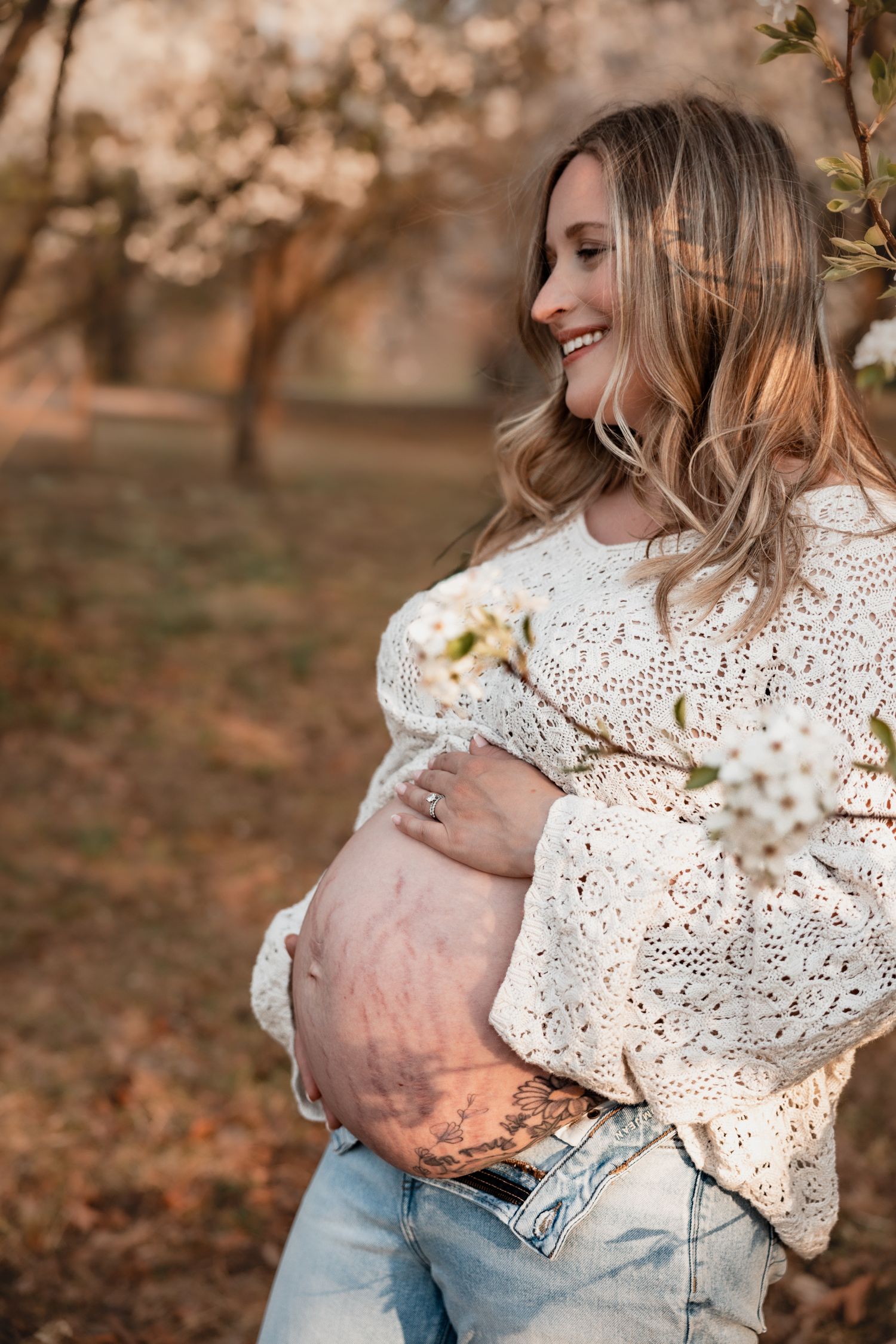 Smiling pregnant woman in white crochet sweater and jeans cradling her bare baby bump outdoors near blooming spring trees.