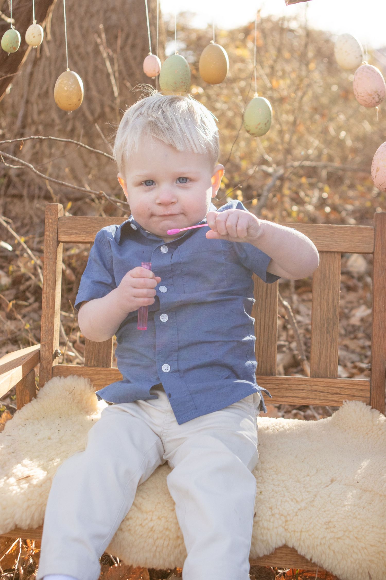 Young child in blue shirt sitting on sheepskin-covered bench with Easter eggs hanging overhead.