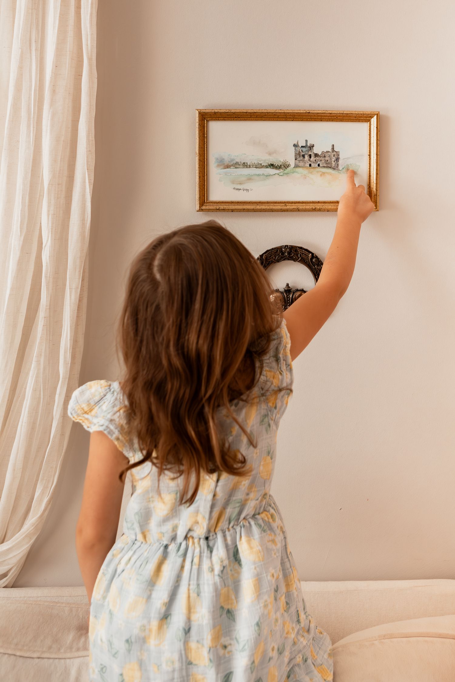 Child in blue floral dress reaching up to point at framed artwork on beige wall.