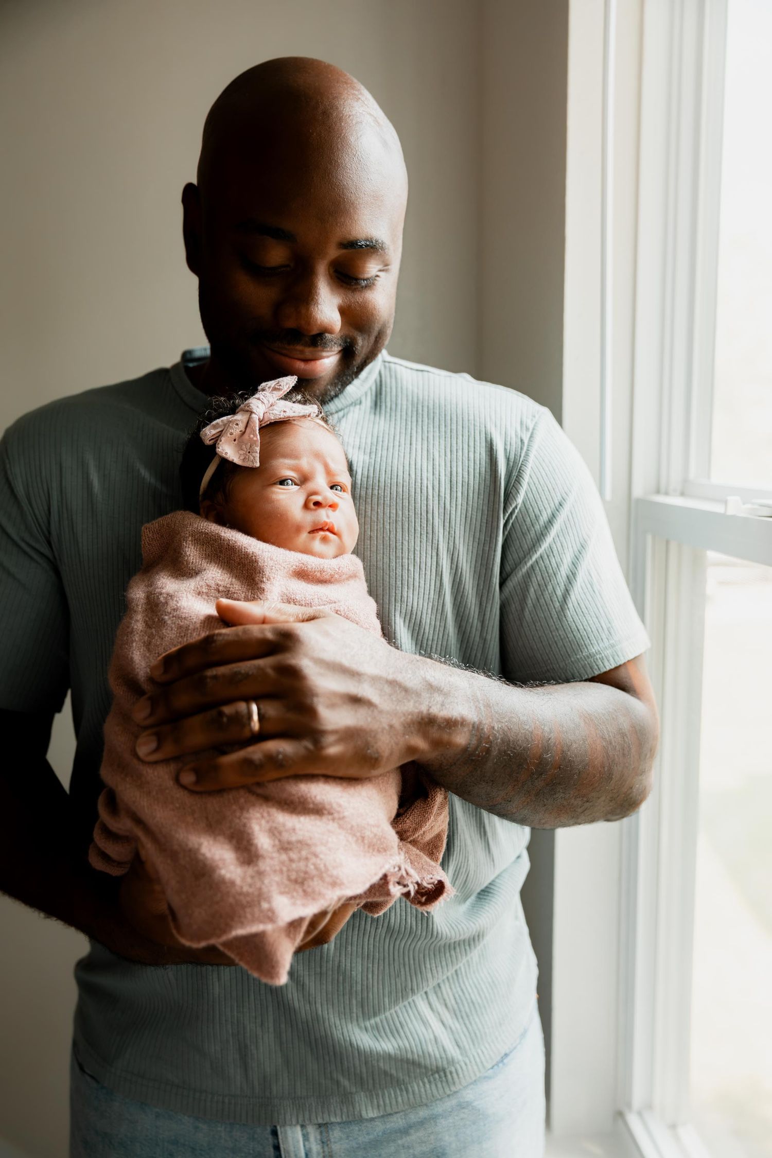 A loving father holds his sleeping newborn baby girl wrapped in a pink blanket near a bright window.