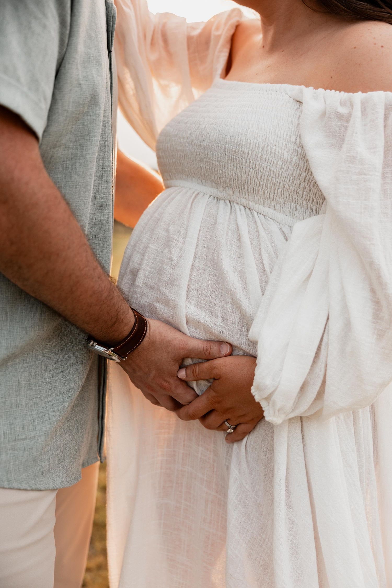 Couple holding hands during maternity photo shoot, pregnant woman in white off-shoulder dress with smocked bodice.