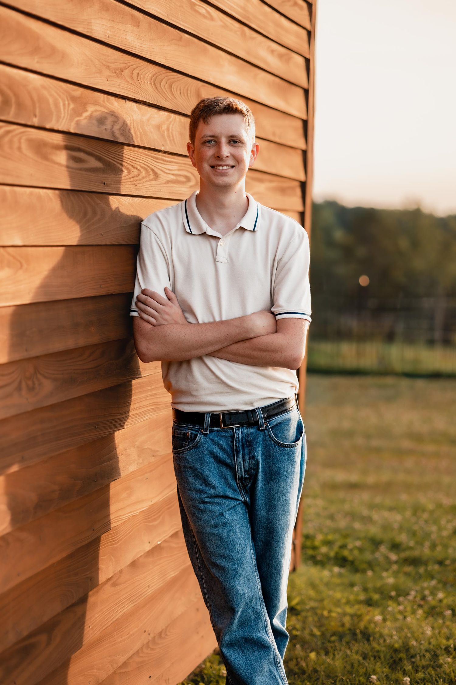 Smiling young man in white polo shirt and jeans leaning against wooden wall in golden hour sunlight.