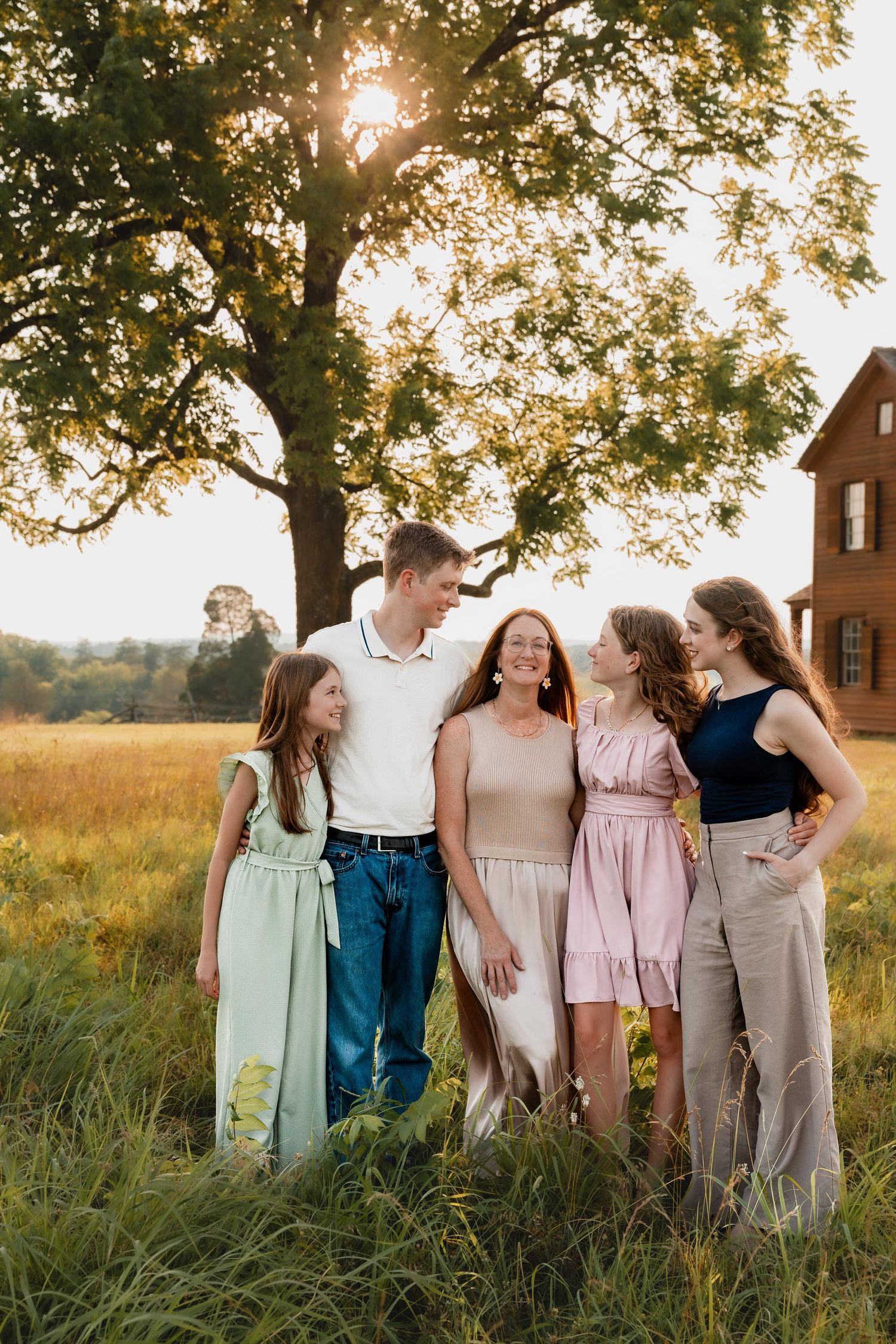 A family of five shares a joyful moment in a golden field near a large tree and red barn.