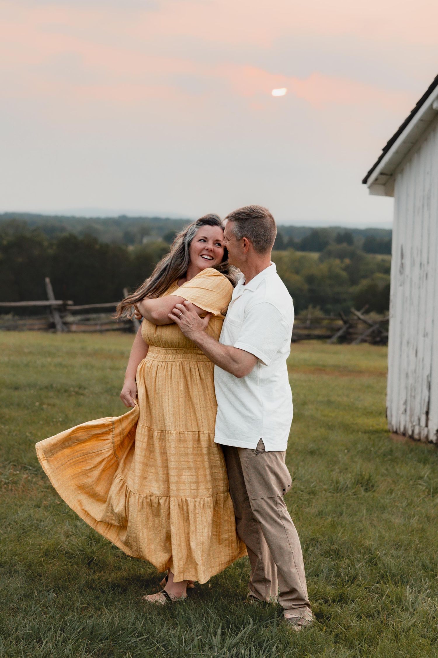 A couple dances romantically at sunset in a field, woman in yellow dress, man in white shirt, near a rustic barn.