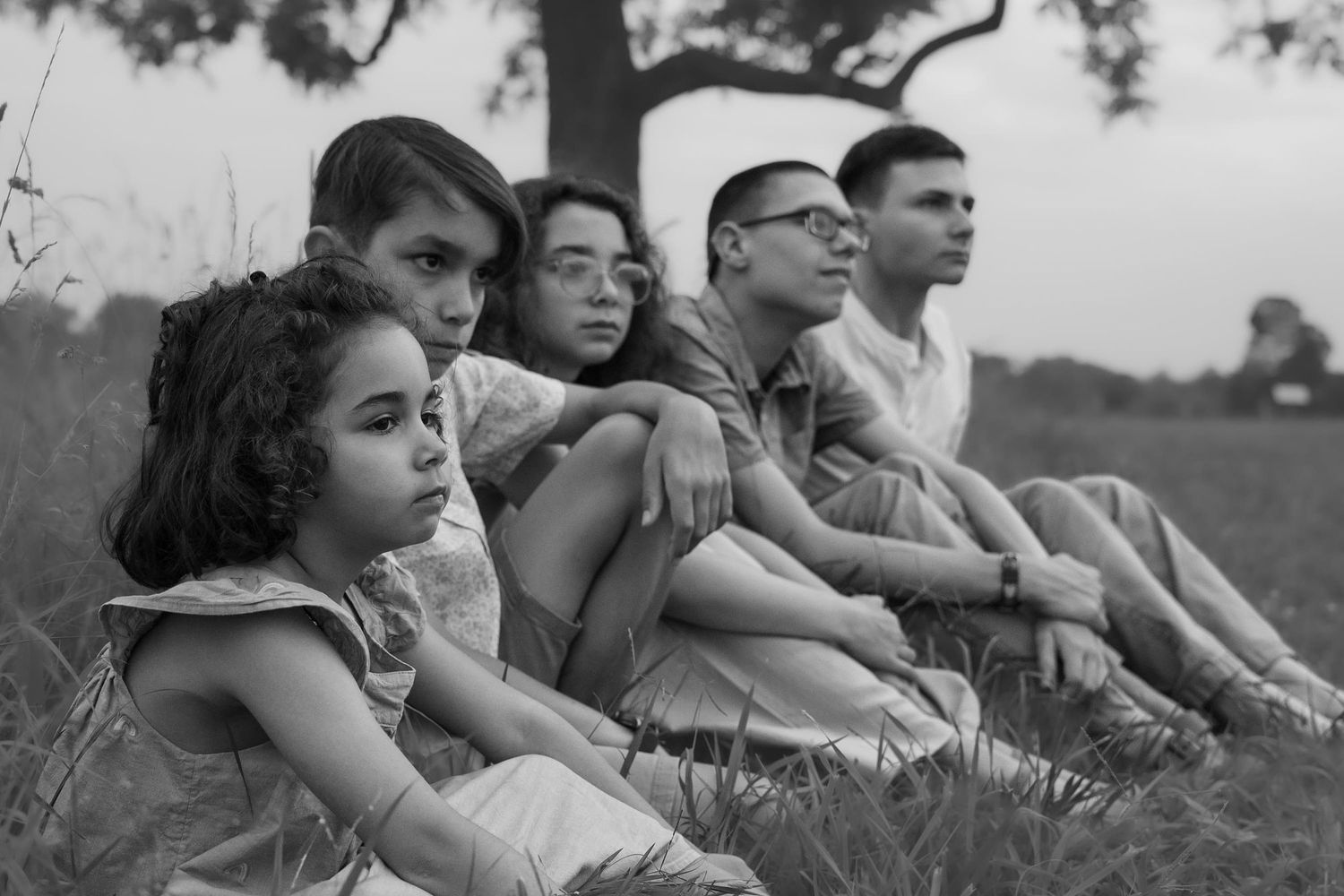 Black and white photo of five people sitting together on grass near a tree in a peaceful outdoor setting.