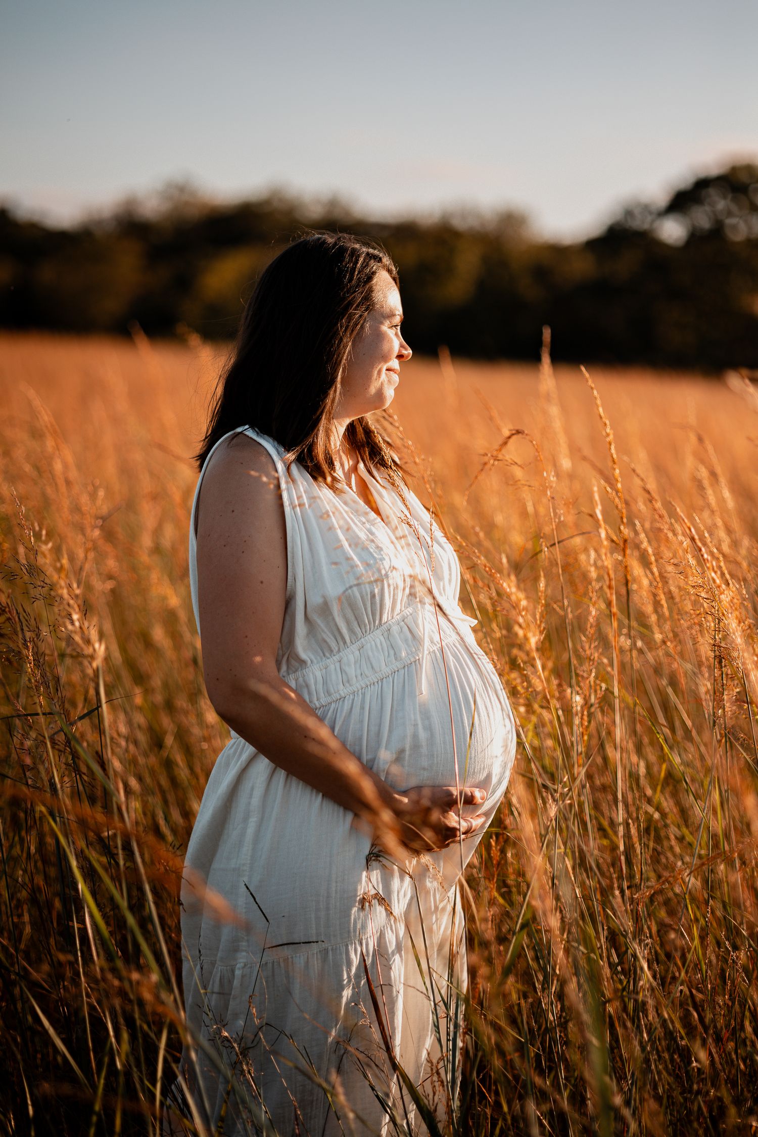 Pregnant woman in white dress standing in golden field at sunset, cradling her baby bump.