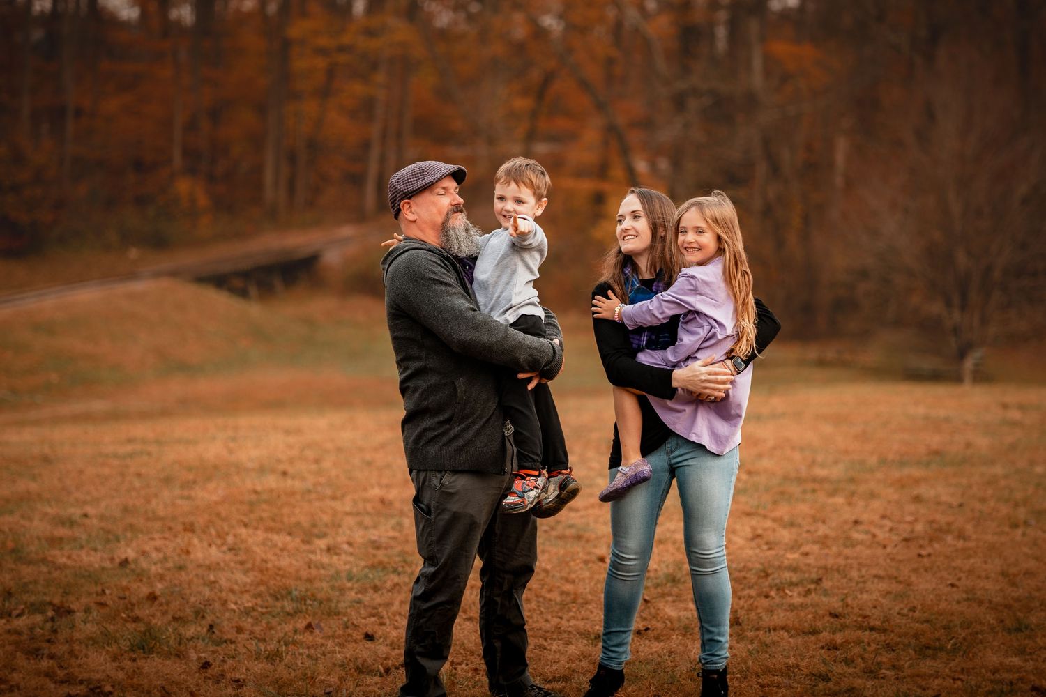 A family of four enjoys a warm autumn day together in a field with orange and brown fall foliage in the background.