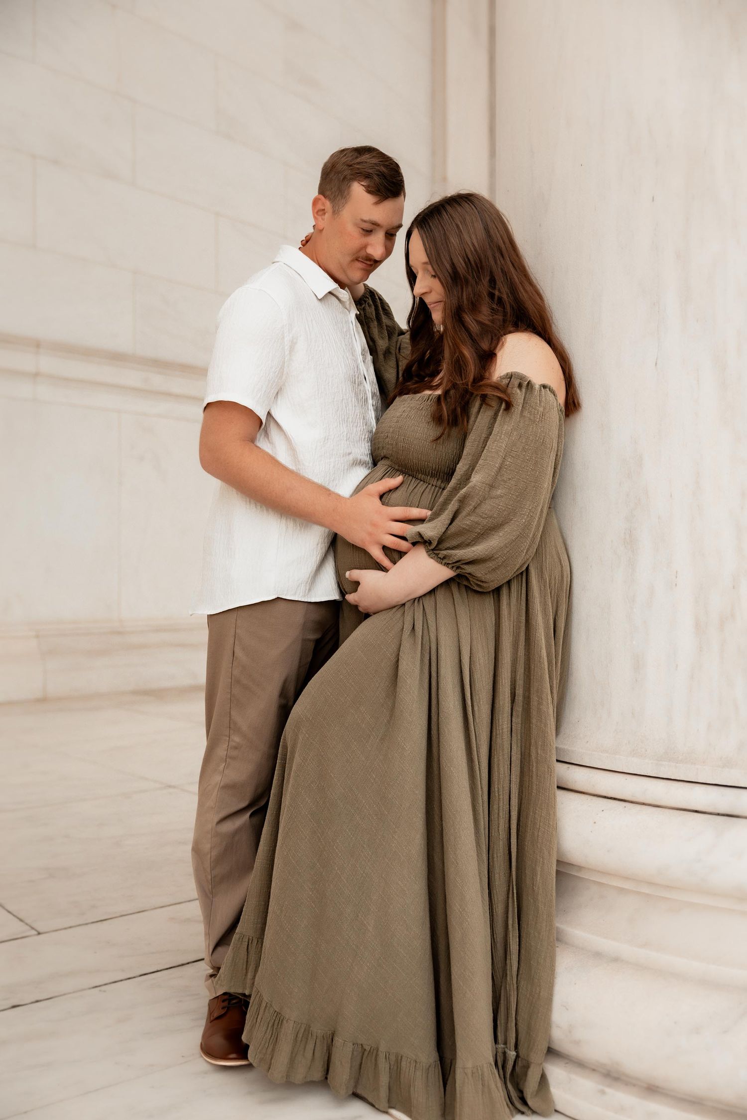 Couple sharing a tender moment during a maternity photo shoot beside marble columns, woman in olive green dress.