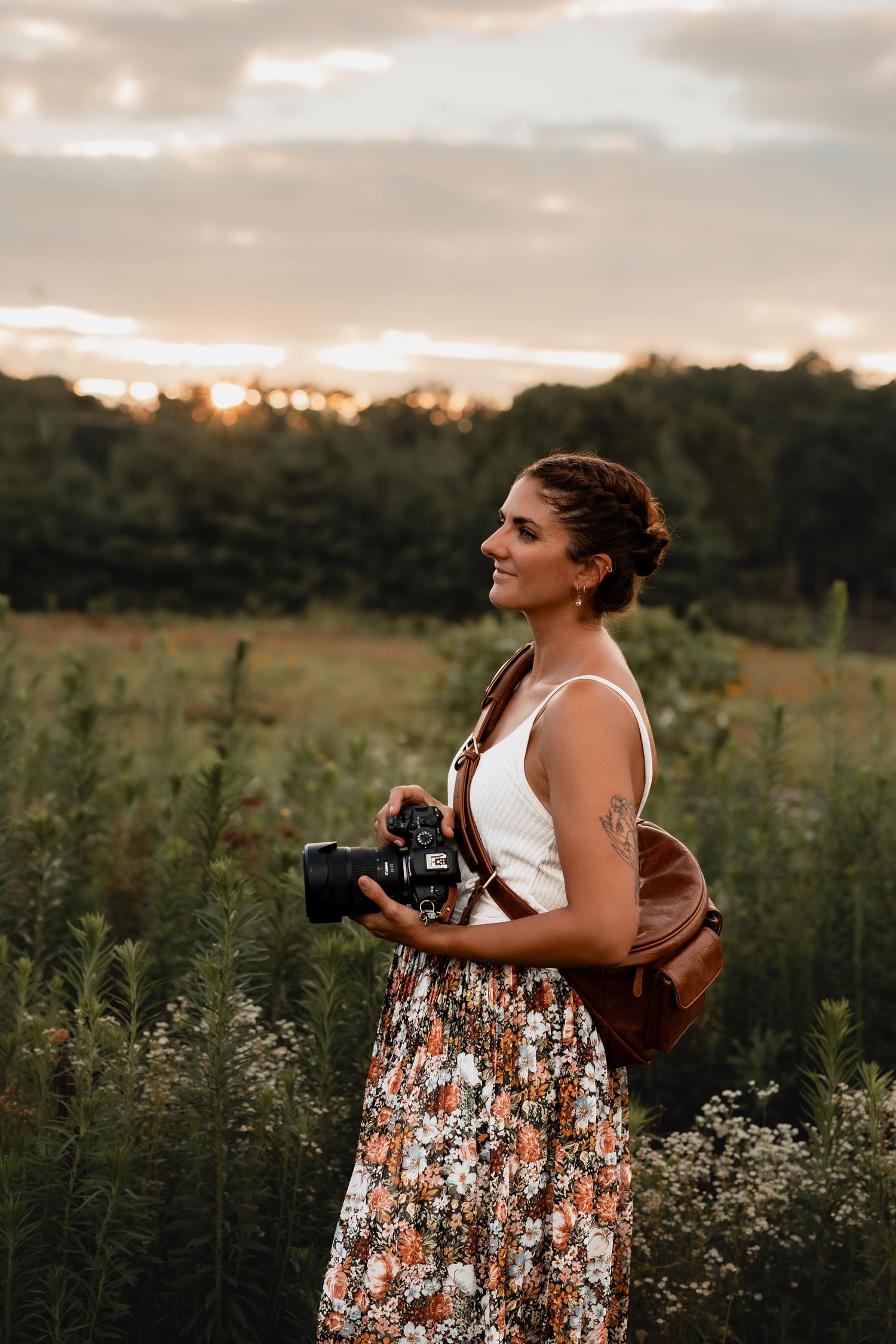 Woman photographer holding DSLR camera in floral skirt and white top standing in a field at golden hour sunset.