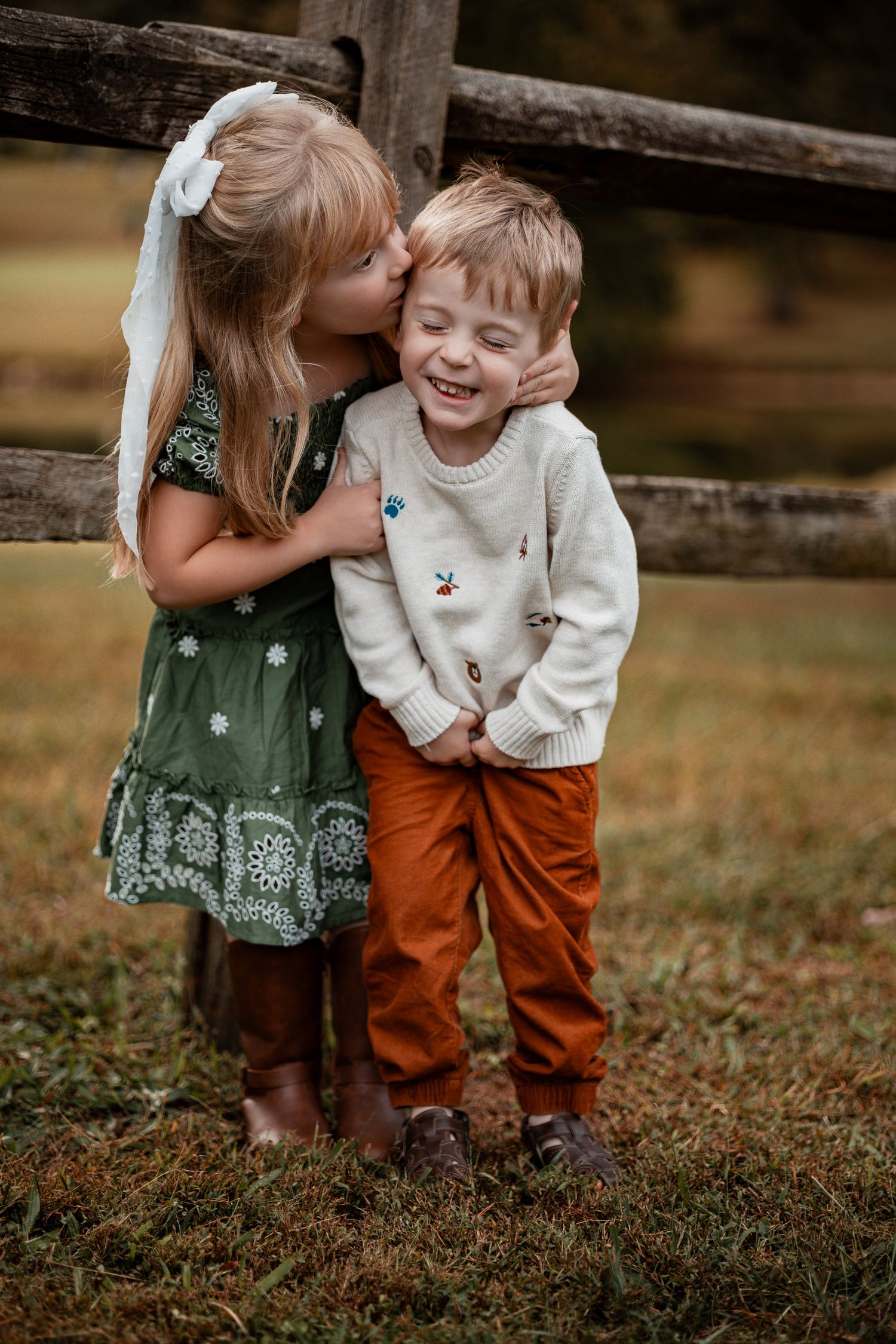 Two young children share a sweet moment together by a wooden fence in autumn, wearing a green dress and rust-colored pants.