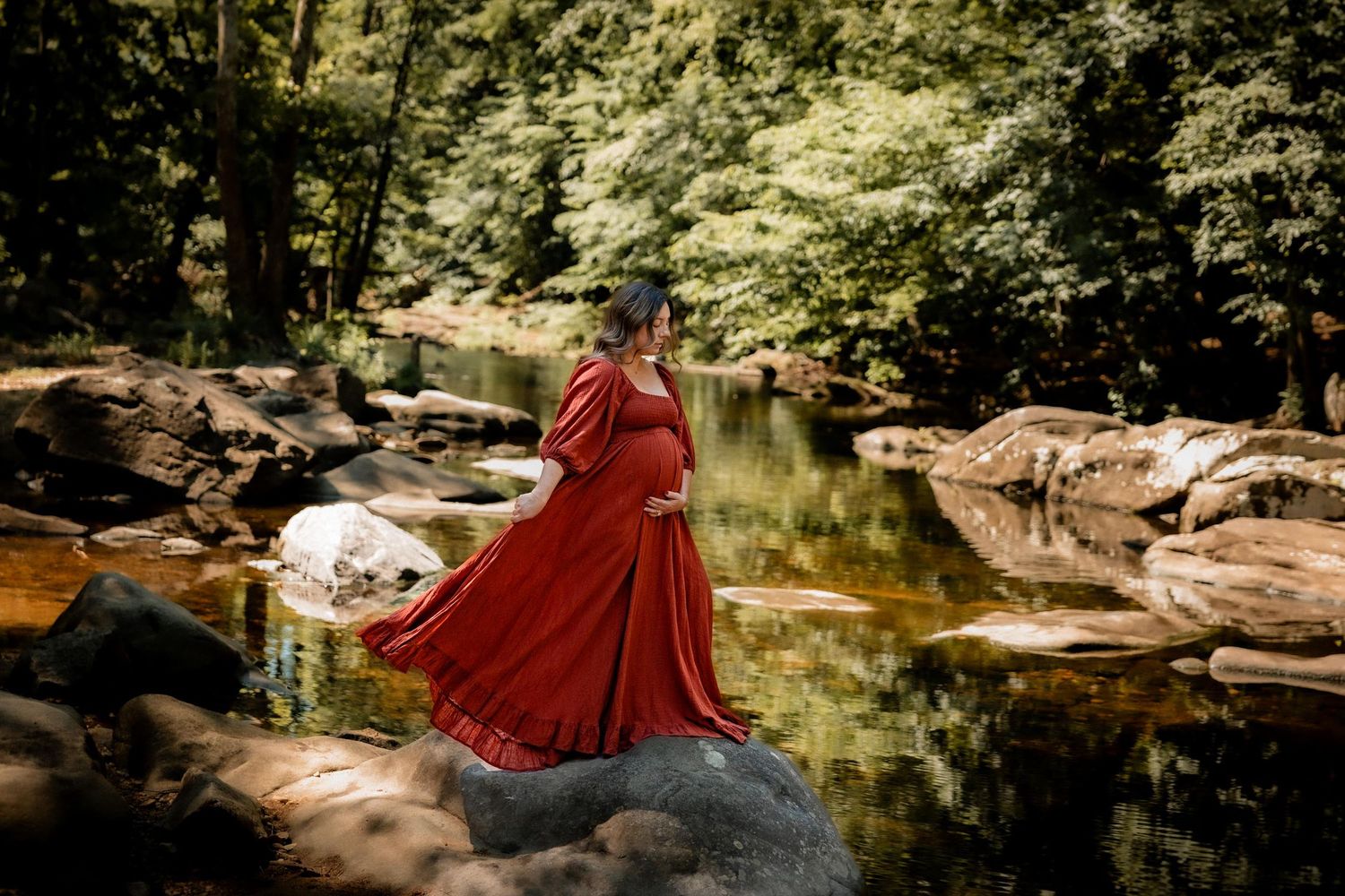 Pregnant woman in flowing red gown standing on rocks by a serene forest creek, maternity photography.