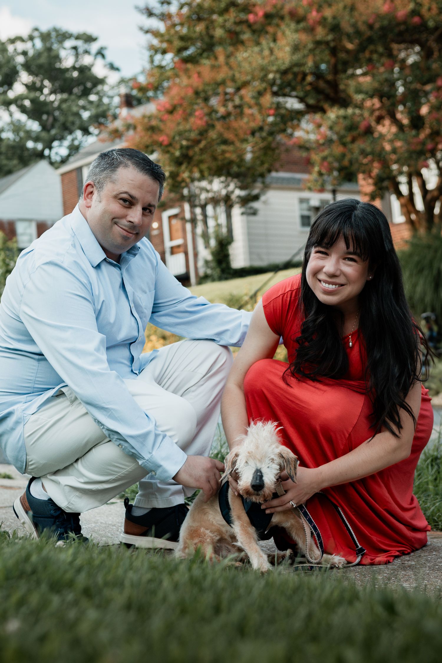 Couple in blue shirt and red dress relaxing on grass with small white dog during autumn outdoor photoshoot.