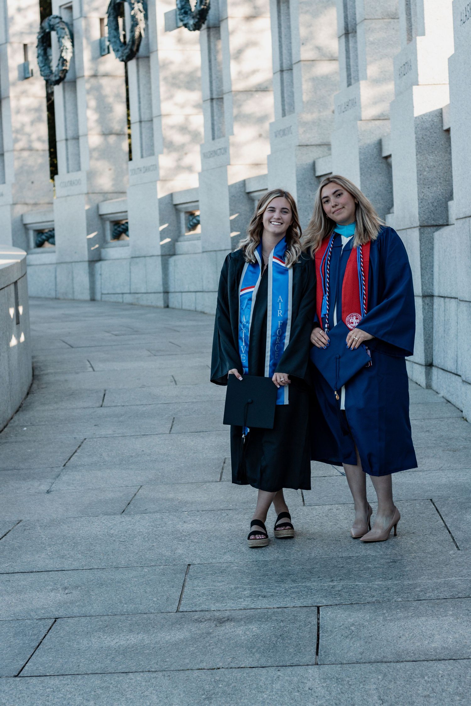 Two graduates wearing navy blue caps and gowns with colorful stoles stand together by stone columns at graduation ceremony.