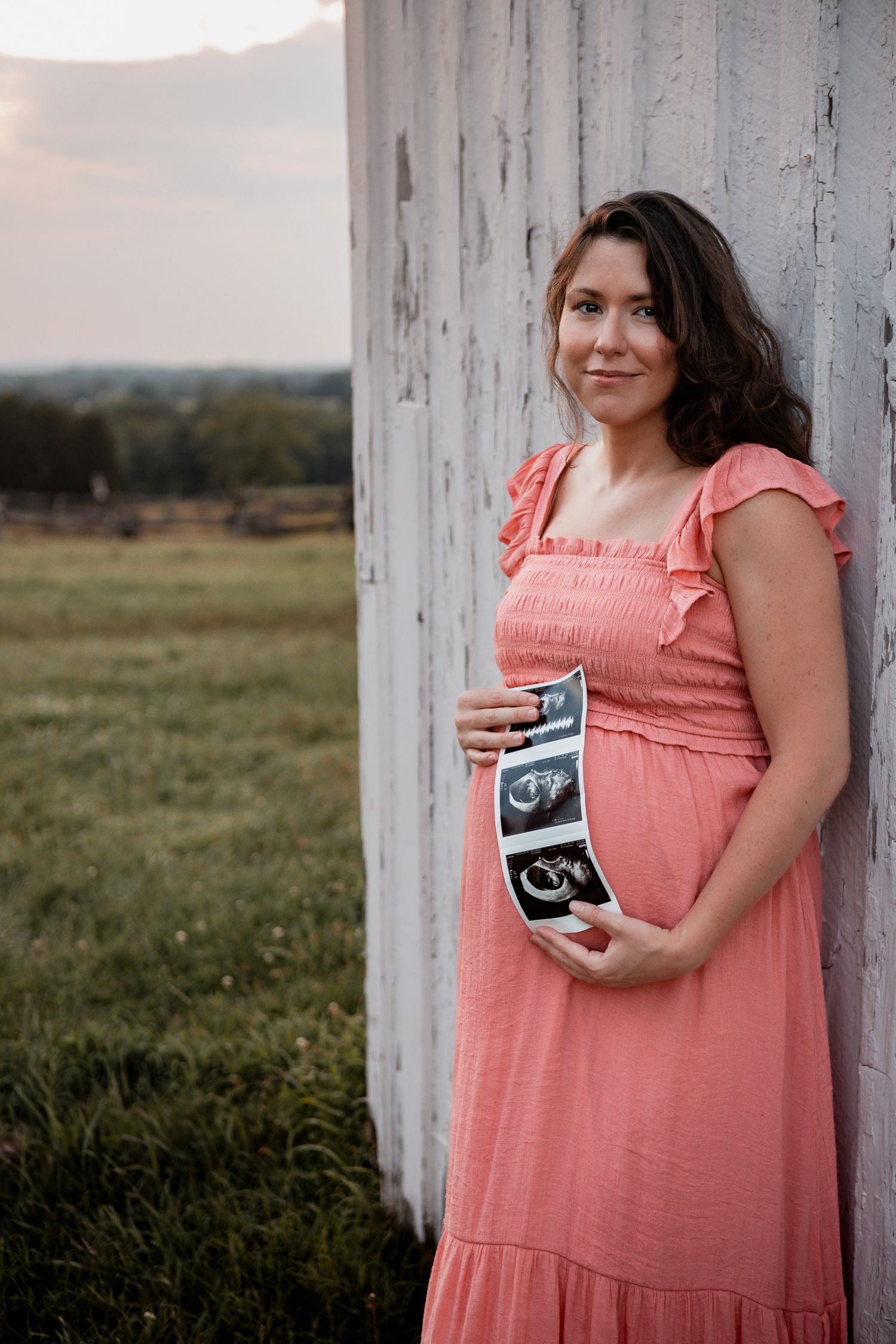 A person in a coral pink dress poses against a white column while holding ultrasound photos in a grassy field at sunset.