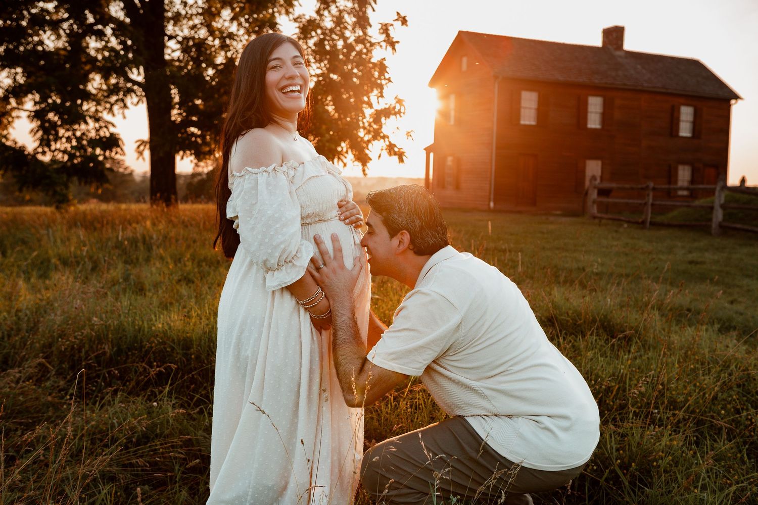Romantic sunset engagement photoshoot in a field with rustic farmhouse silhouette in warm golden light.