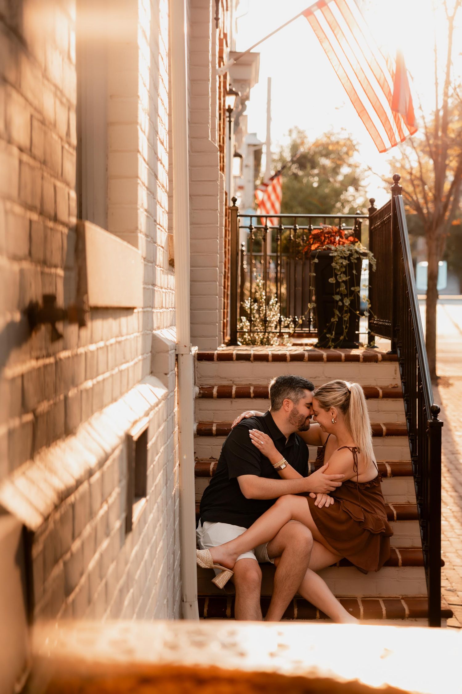 Couple sharing a romantic moment on sunlit brick steps with American flags in the background.