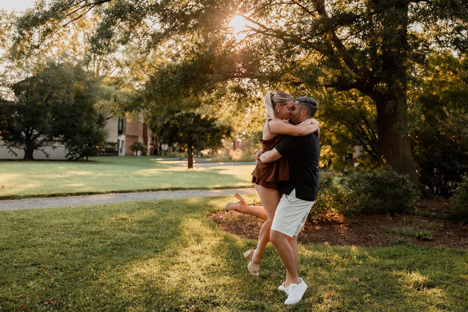 Couple embracing and spinning in a sunlit park during golden hour, surrounded by lush green trees and grass.