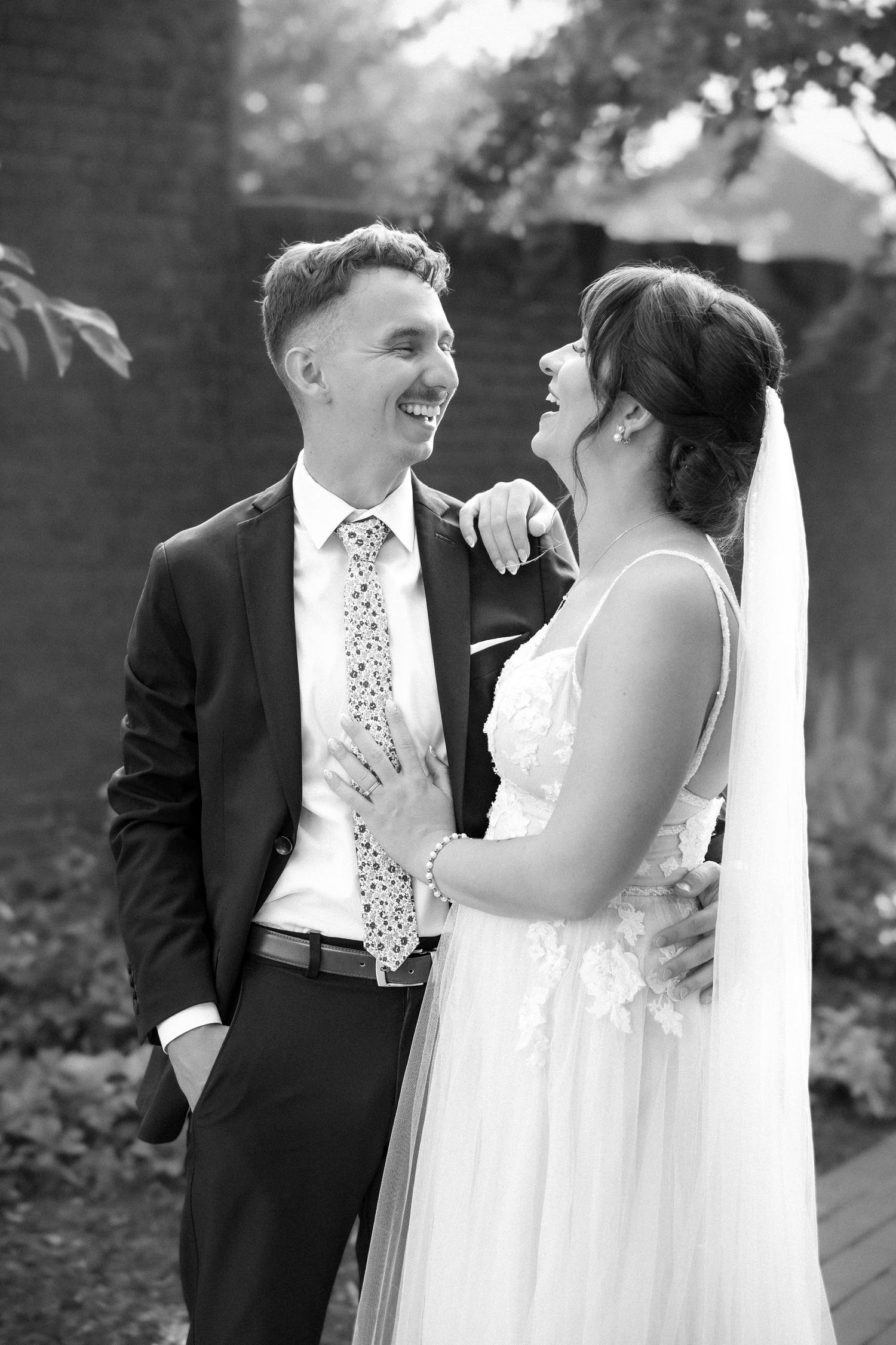 Black and white photo of a laughing bride and groom sharing a joyful moment outdoors on their wedding day.