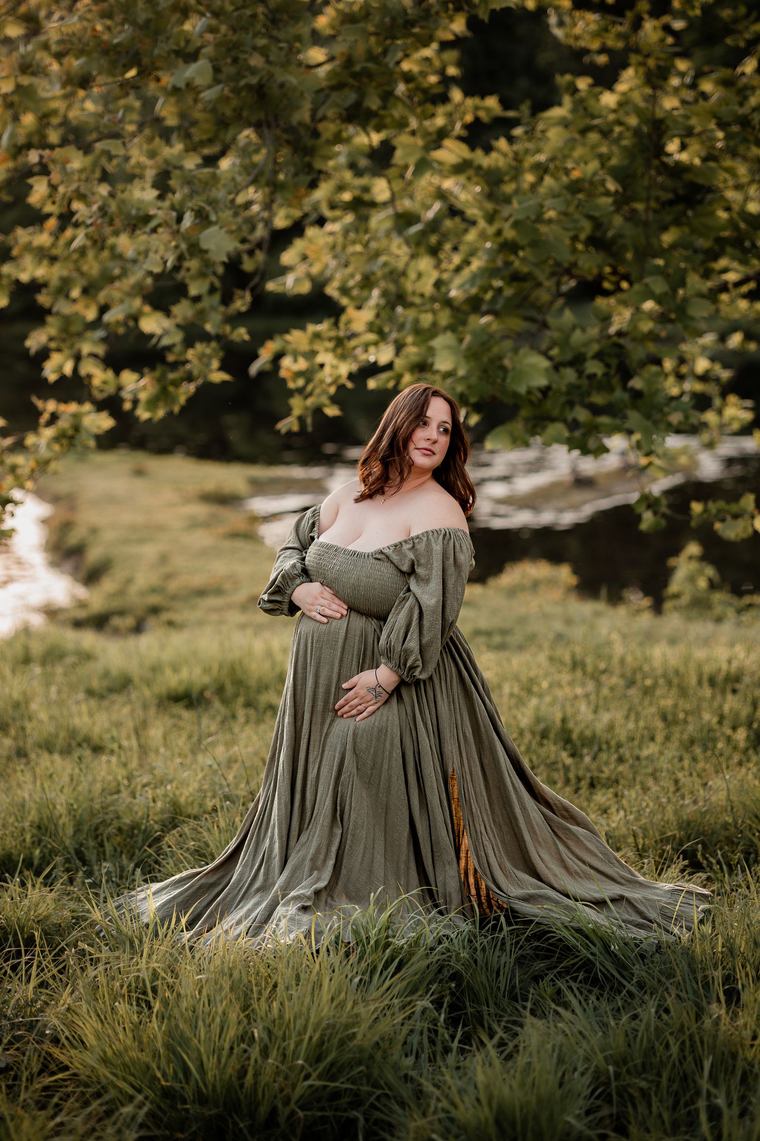 Pregnant woman in olive green off-shoulder gown posing outdoors in lush green field with trees.