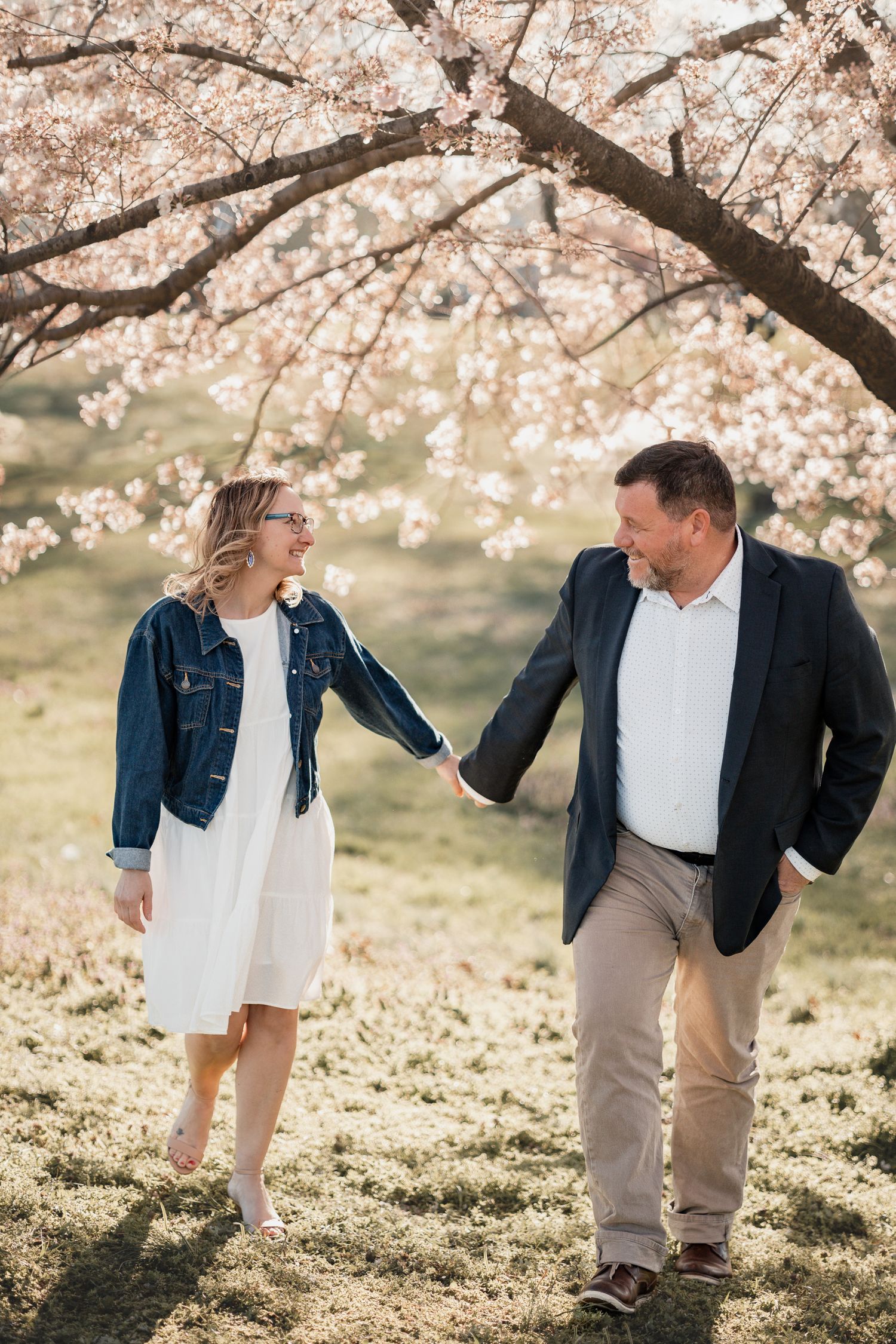 Couple holding hands and smiling under blooming cherry blossom trees in spring, woman in denim jacket and white dress.