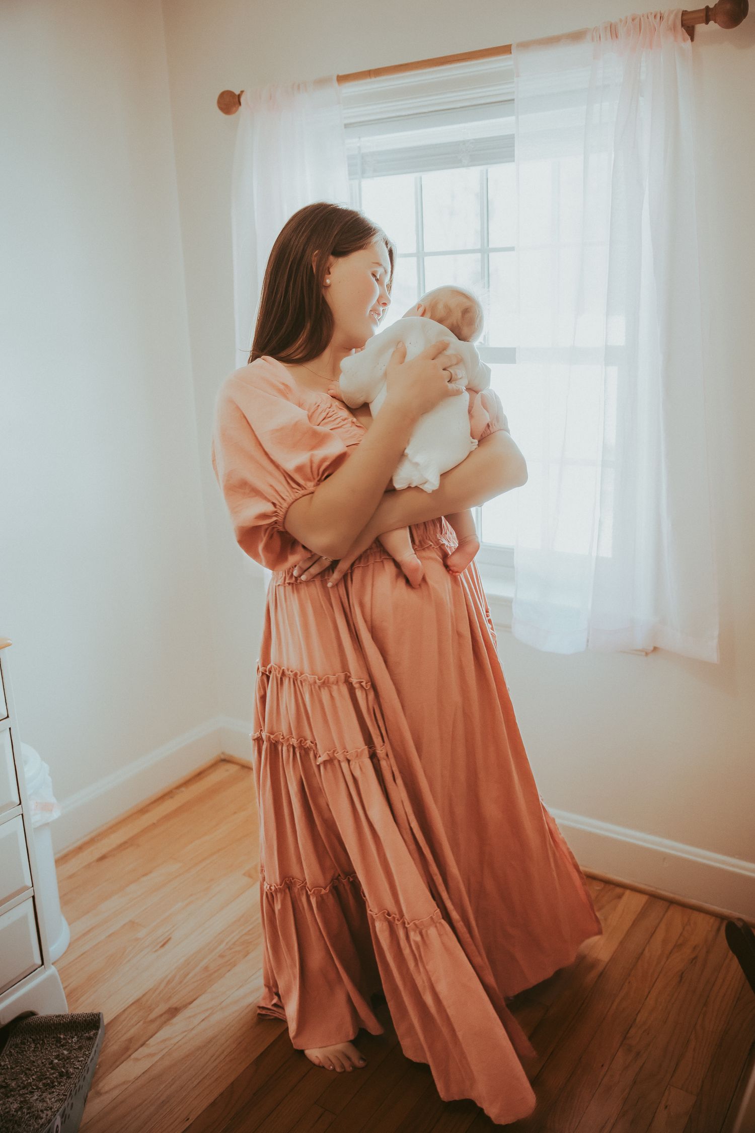 A woman in a terracotta maxi dress holds a newborn baby near a bright window in a nursery.