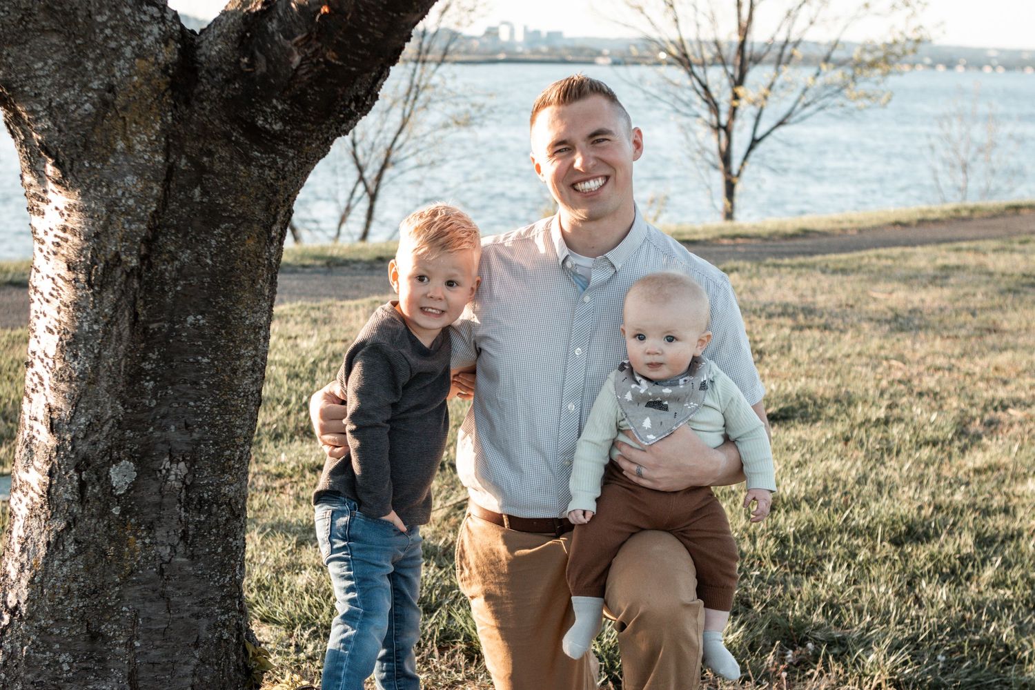 Three people smile together on a scenic autumn day by the waterfront with bare trees in the background.