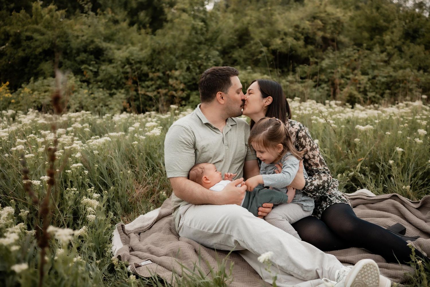 Family of four sits in a wildflower meadow, parents kissing while holding newborn and toddler on a blanket.