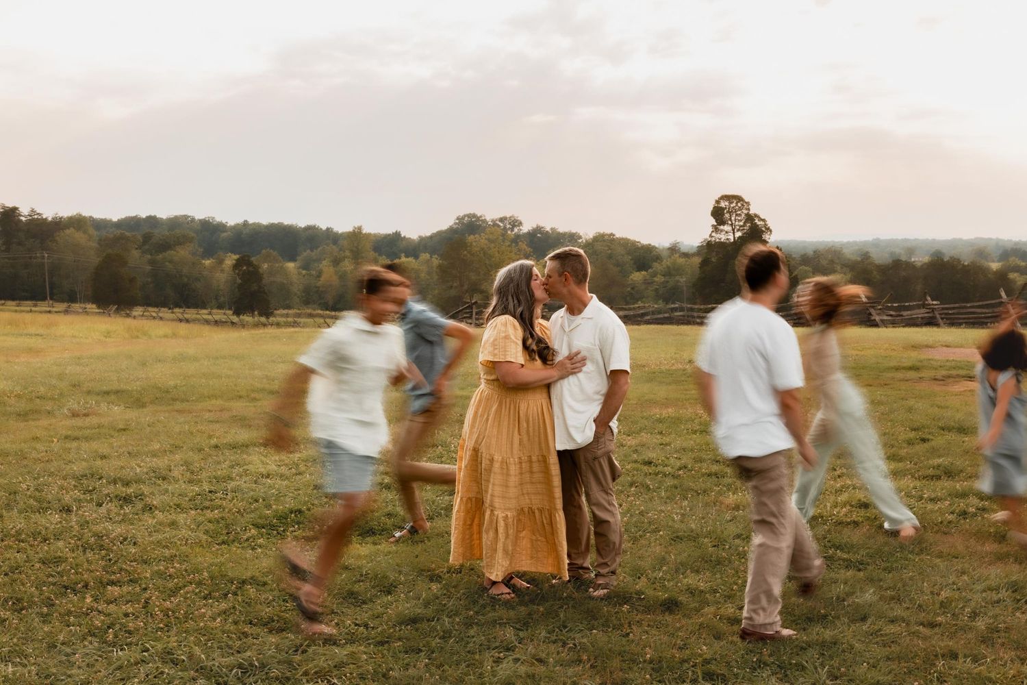 Couple shares a tender kiss surrounded by blurred moving family in a lush green field.