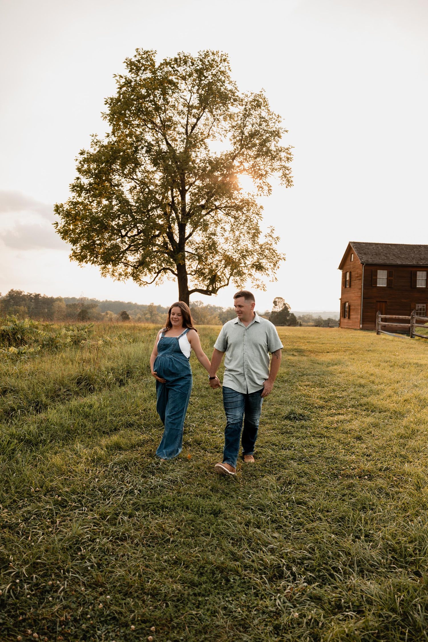 Pregnant couple holding hands walking in golden hour sunlit field with large tree and rustic barn in background.
