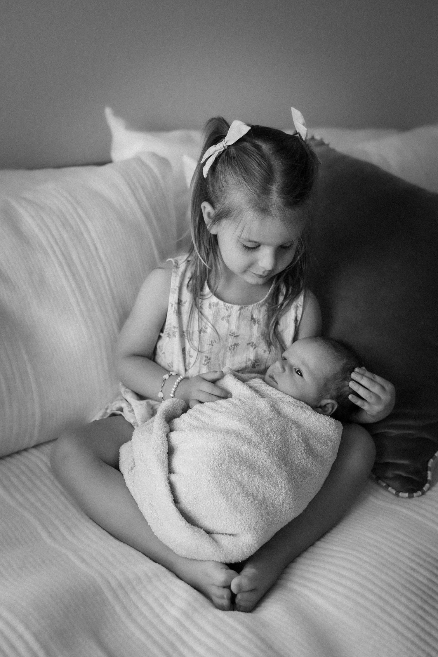 Black and white portrait of a young girl tenderly holding a swaddled newborn baby while seated on a bed.