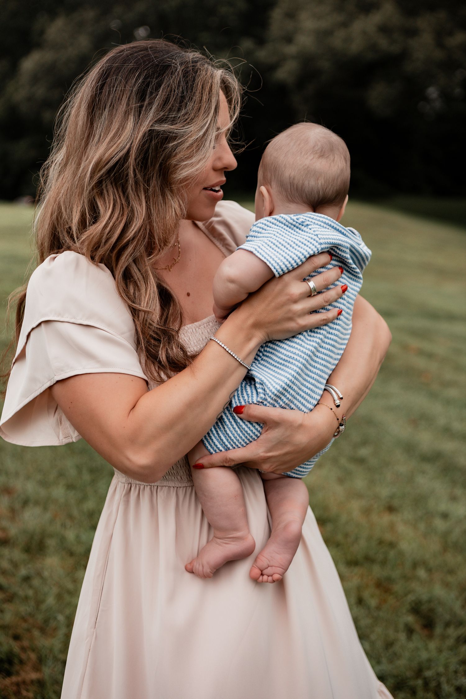 A mom wearing a blush dress embraces and plays with her baby in blue stripes outdoors in a park setting.