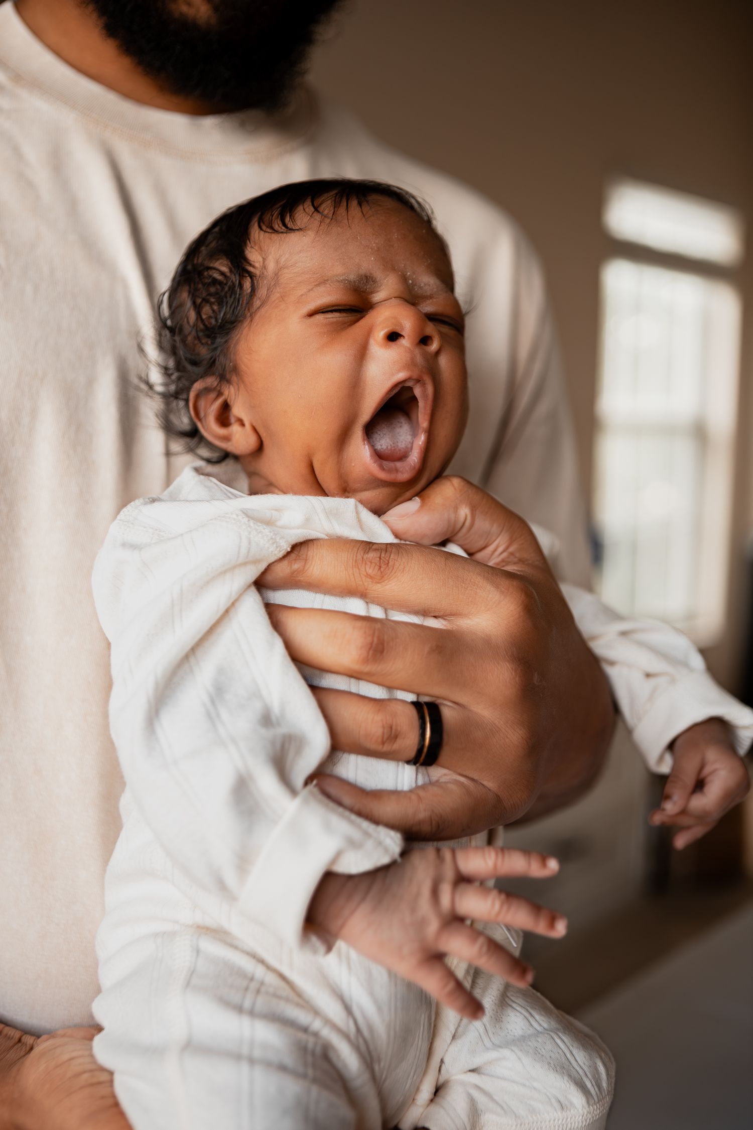 Baby in white onesie yawns while being held against soft background.