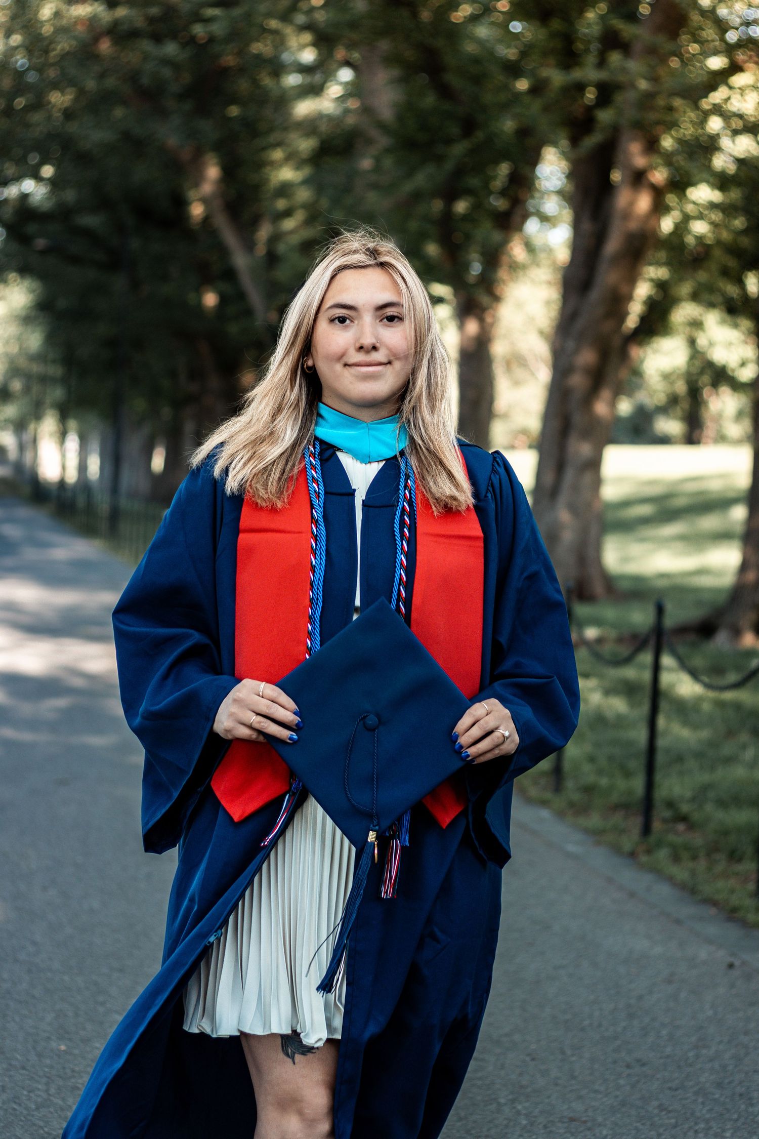 Graduate in blue cap and gown with red stole poses for photos in a park setting.